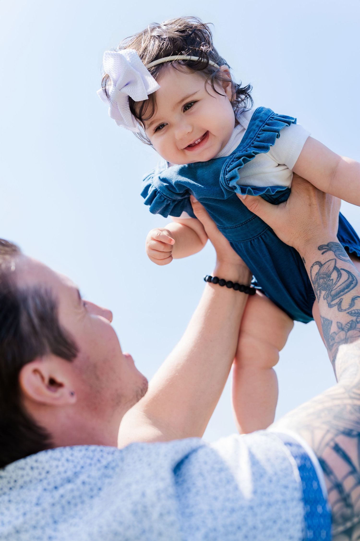 Baby wearing blue outfit and white bow smiles while being lifted in the air.