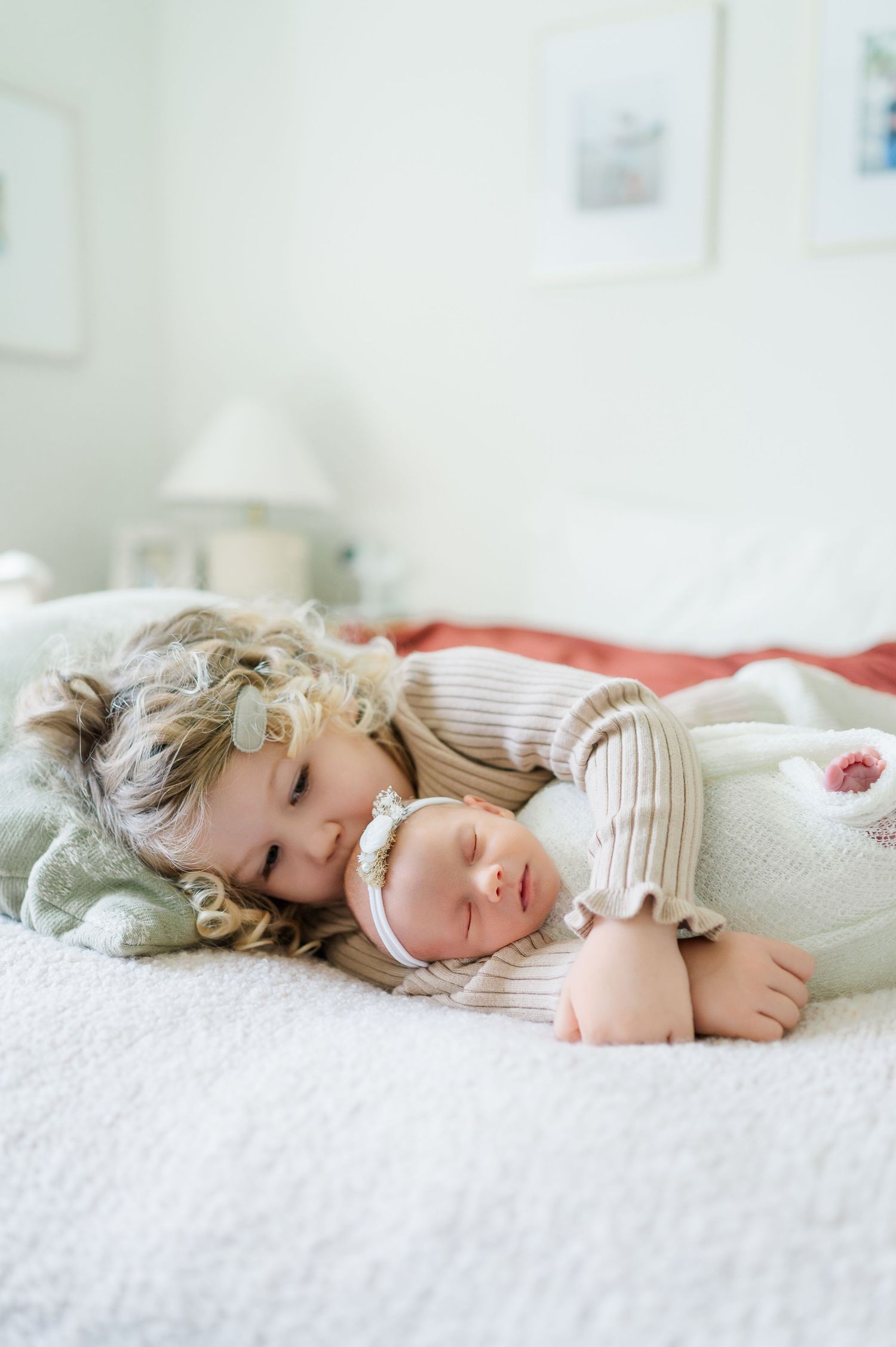 Two siblings wearing cream colored knit sweaters cuddle together while laying on a white bed.