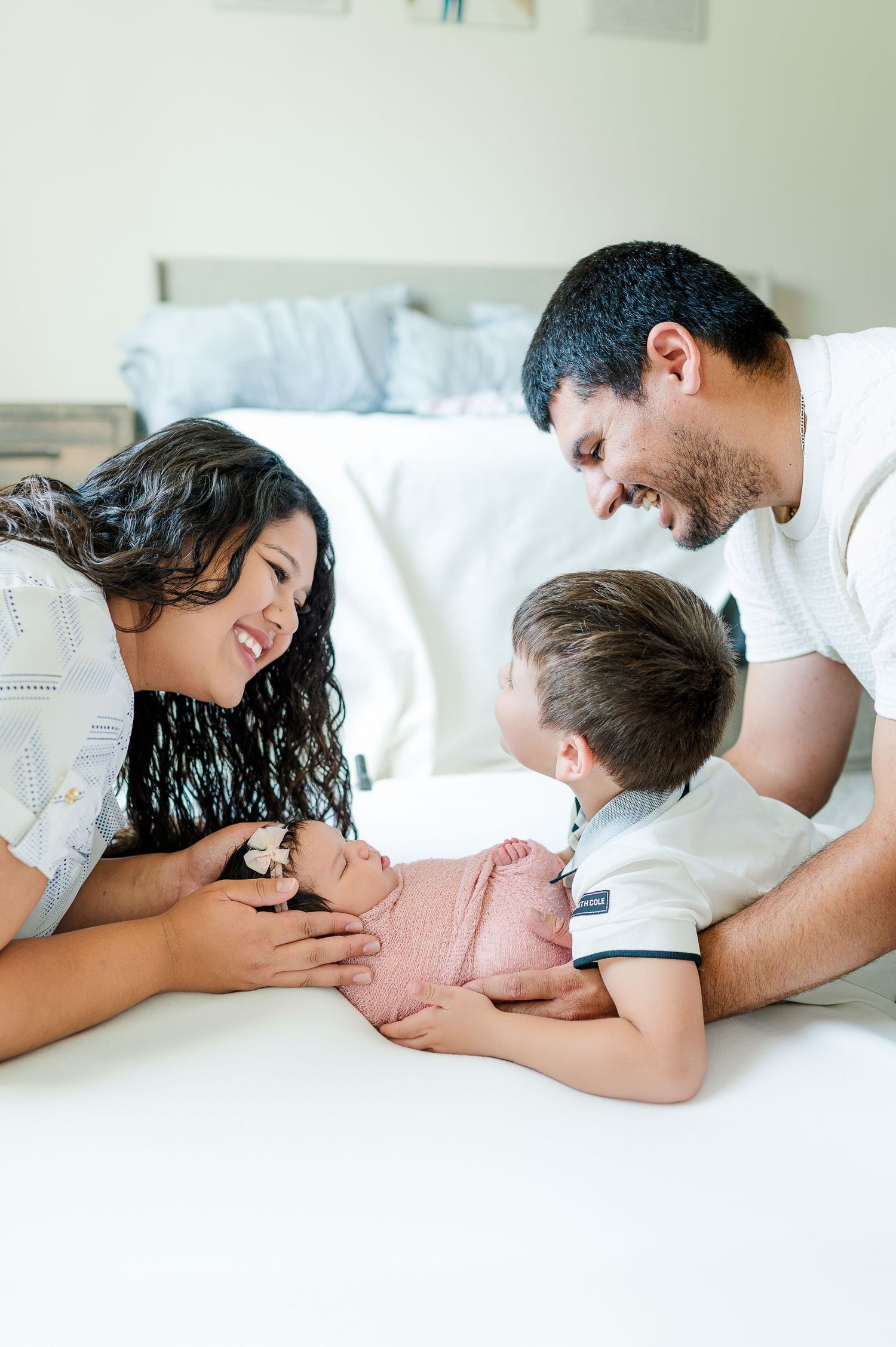 A joyful family of four admires their newborn baby wrapped in pink on a white bed at home.