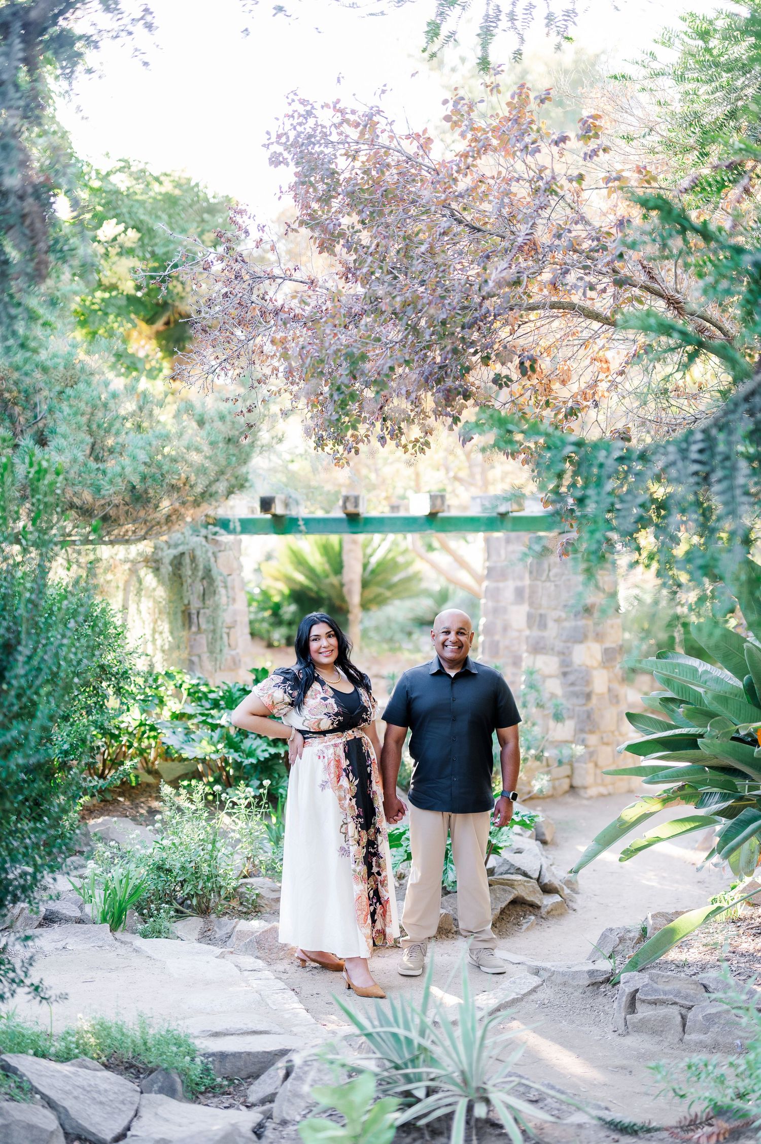 A couple walks together through a lush garden path surrounded by flowering trees and stone walls on a sunny day.