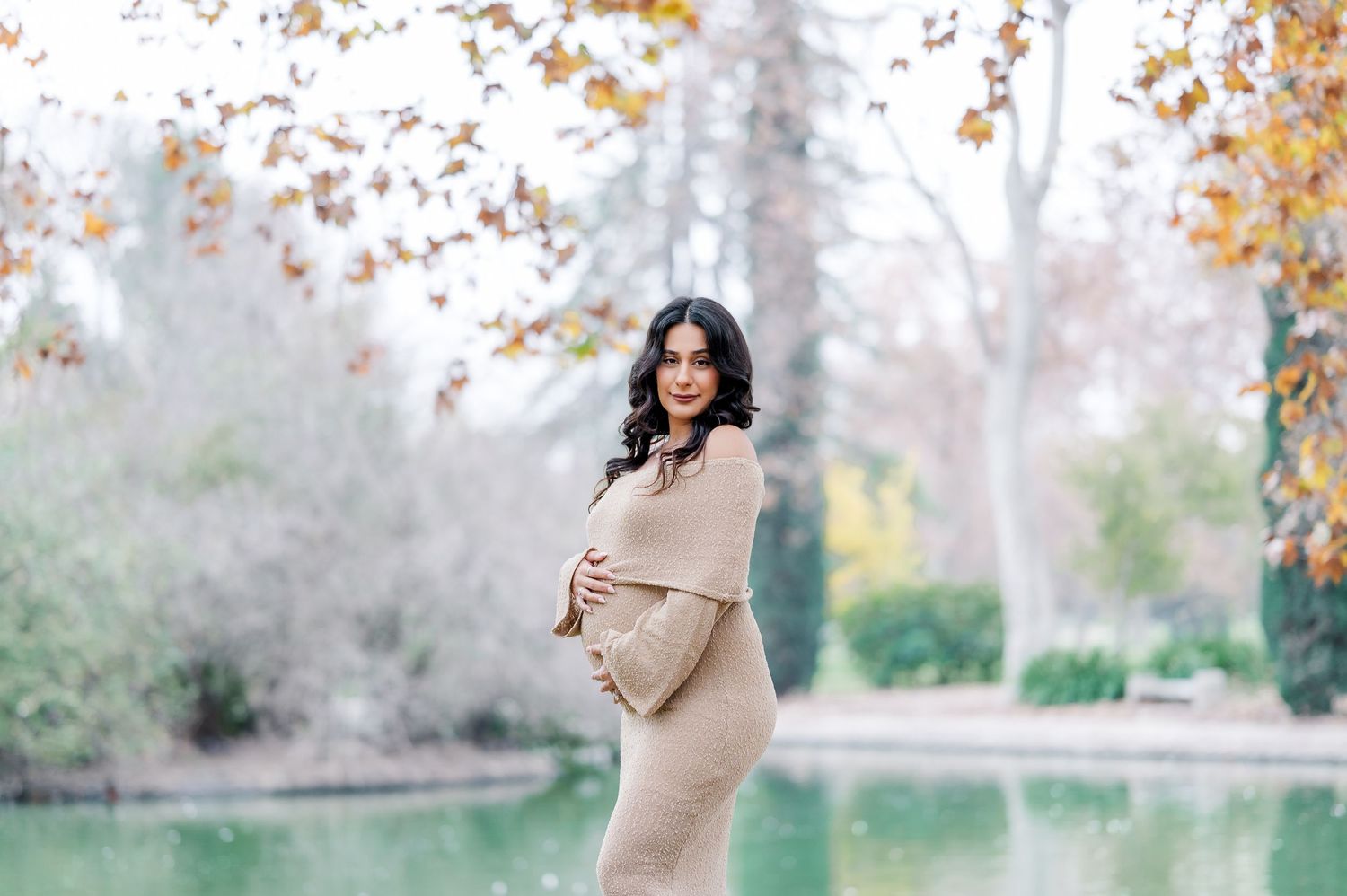 A woman in a beige sweater walks through a park during autumn with golden leaves falling in the background.