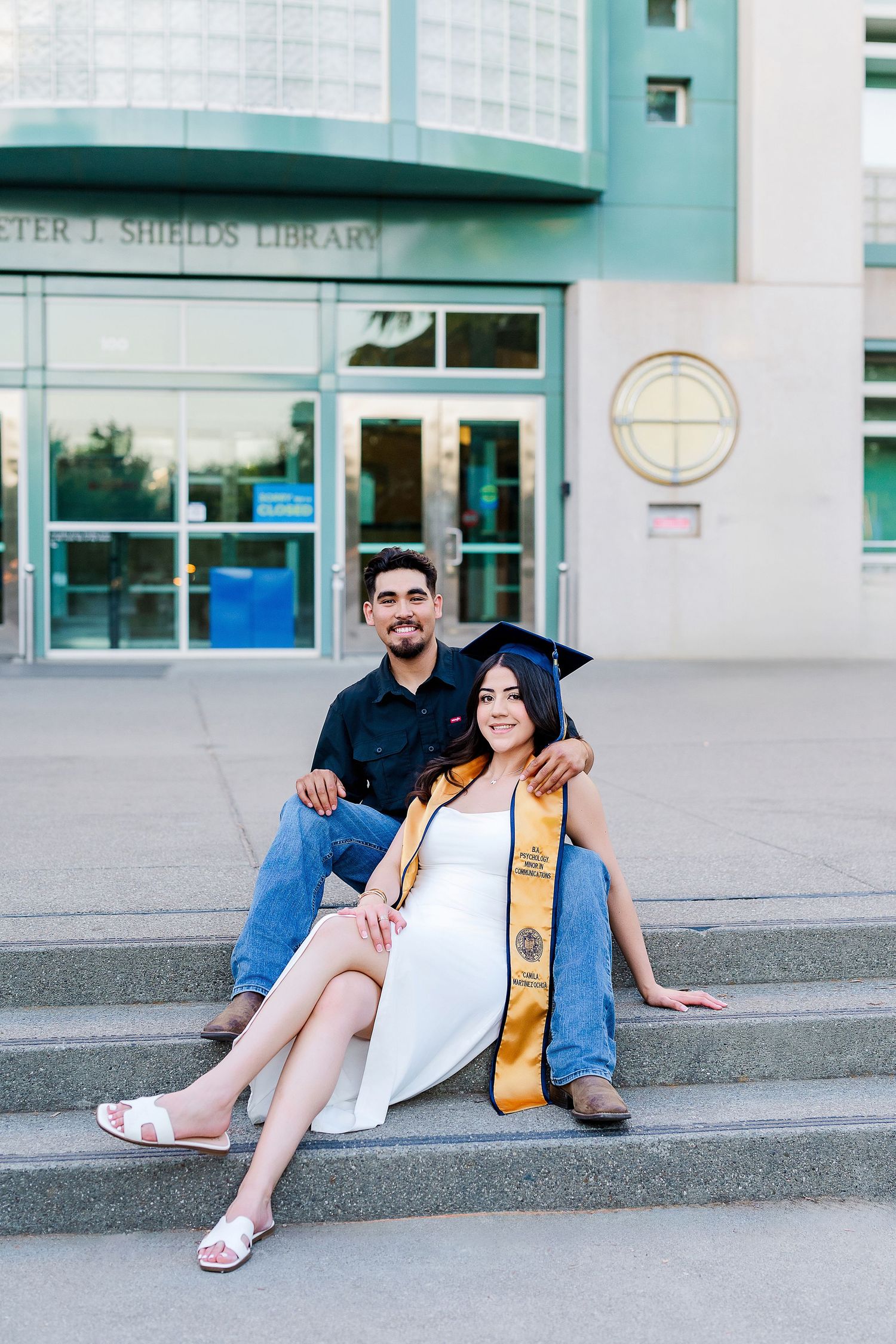 Graduate posing casually on concrete steps outside a modern building with glass windows and teal accents.