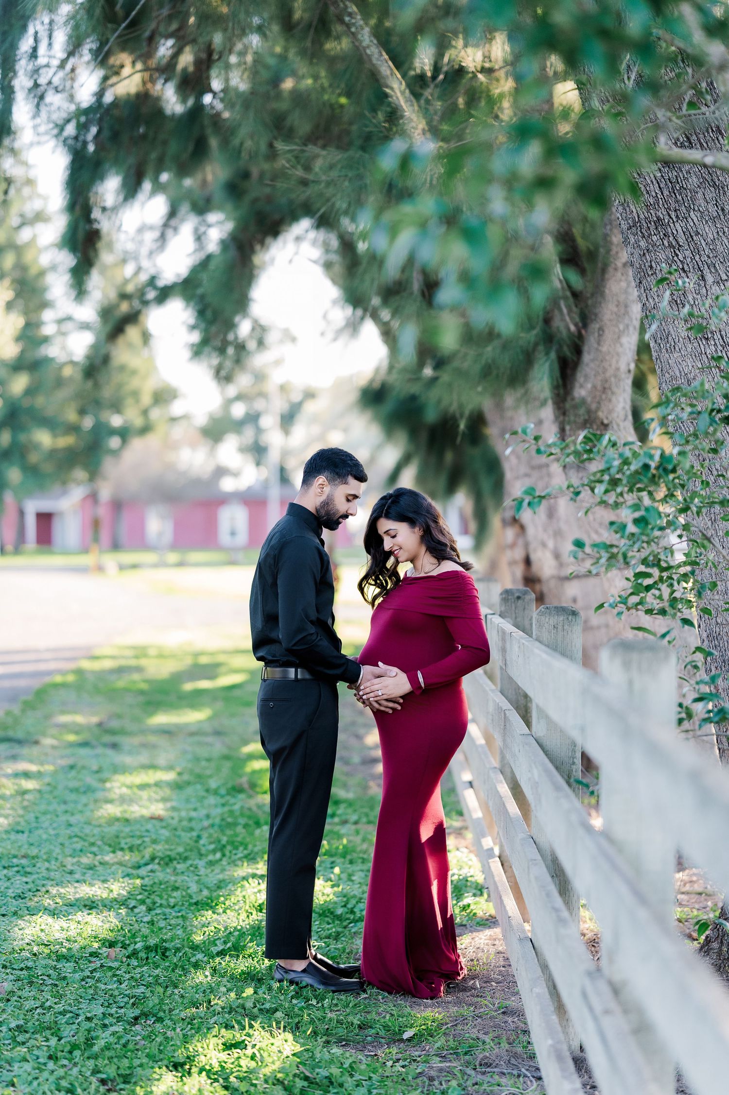 A romantic maternity photoshoot featuring a couple by a white fence with trees in the background.