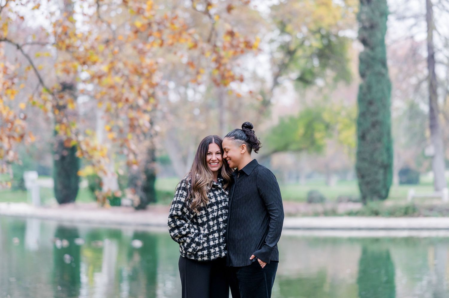 A couple shares tender moments while walking through a park with autumn trees and fallen leaves in the background.