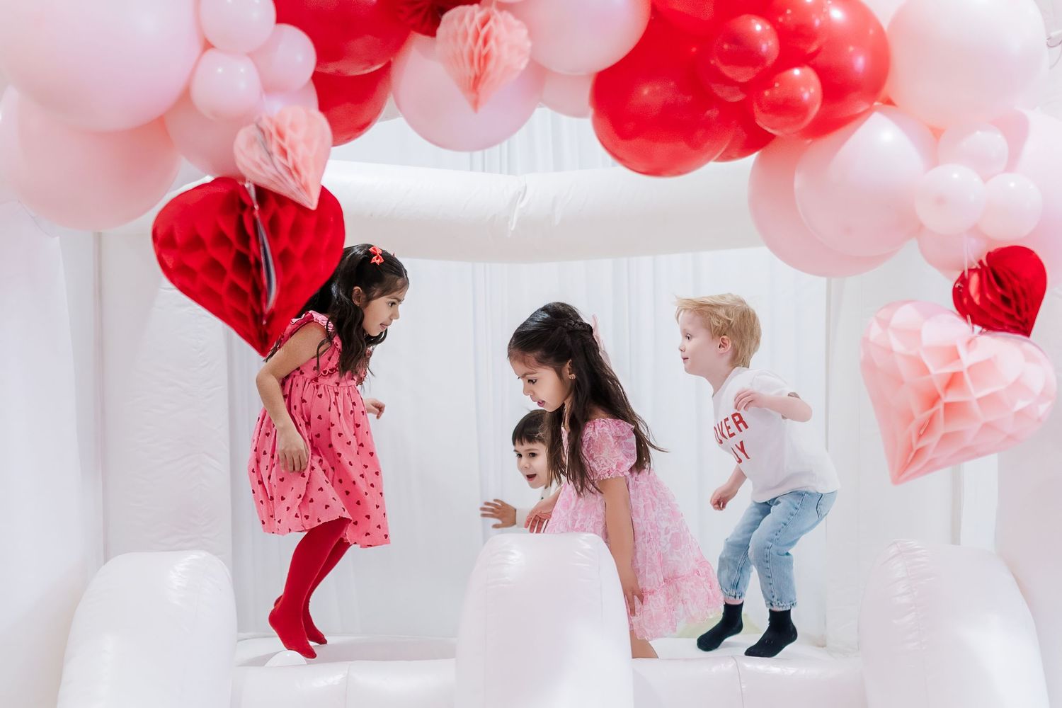 Children playing in a white bounce house decorated with pink and red heart-shaped balloons.