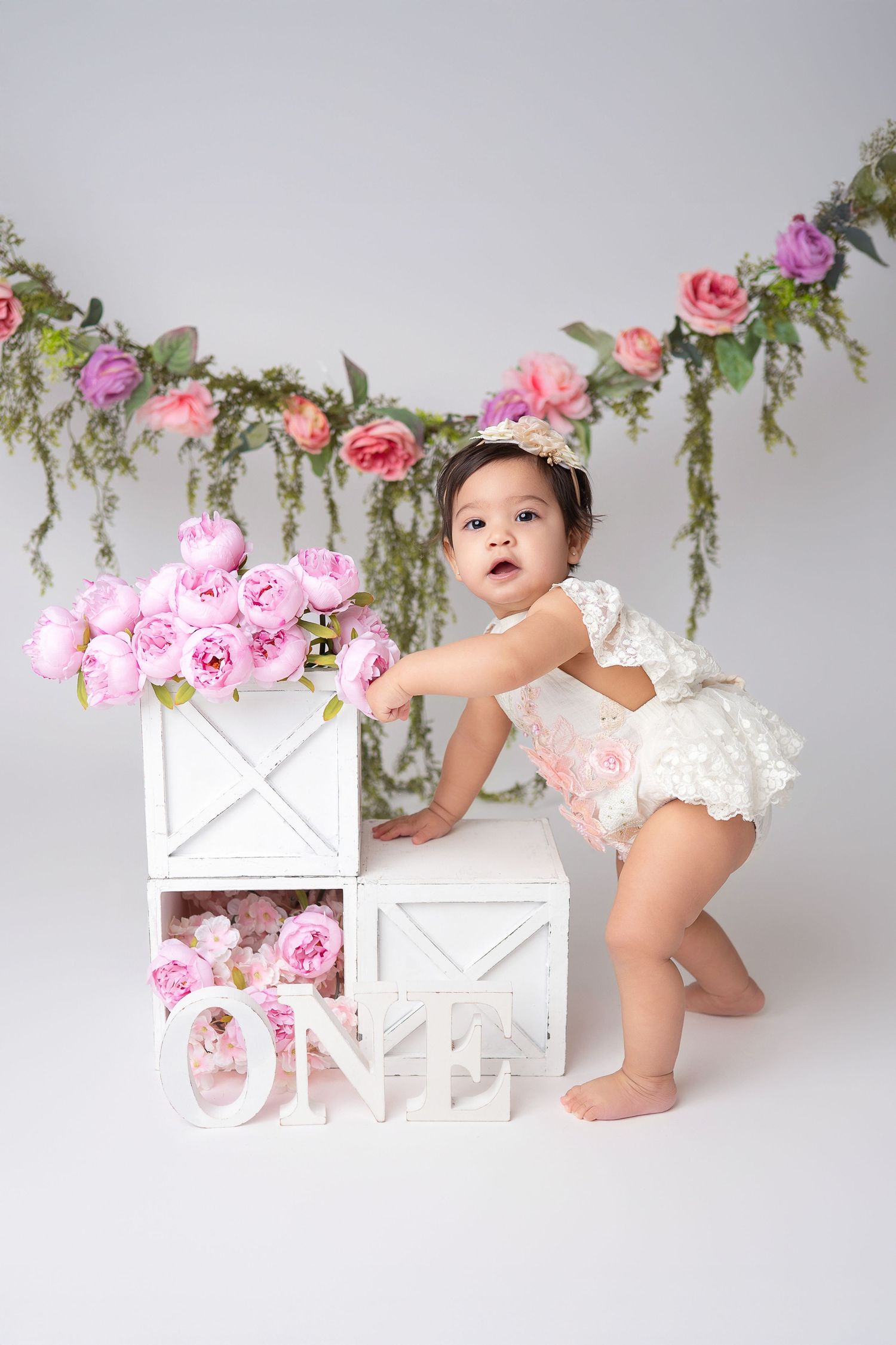 Baby in white outfit next to floral decorations and pink roses during first birthday photo shoot.
