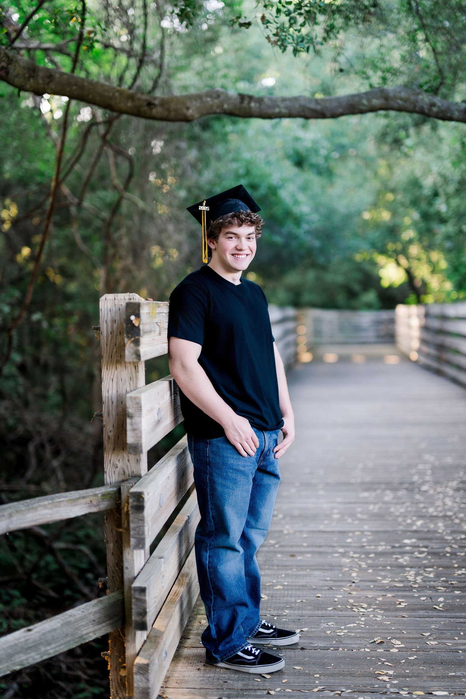 Smiling young man in black t-shirt and graduation cap poses on wooden bridge surrounded by lush green trees.