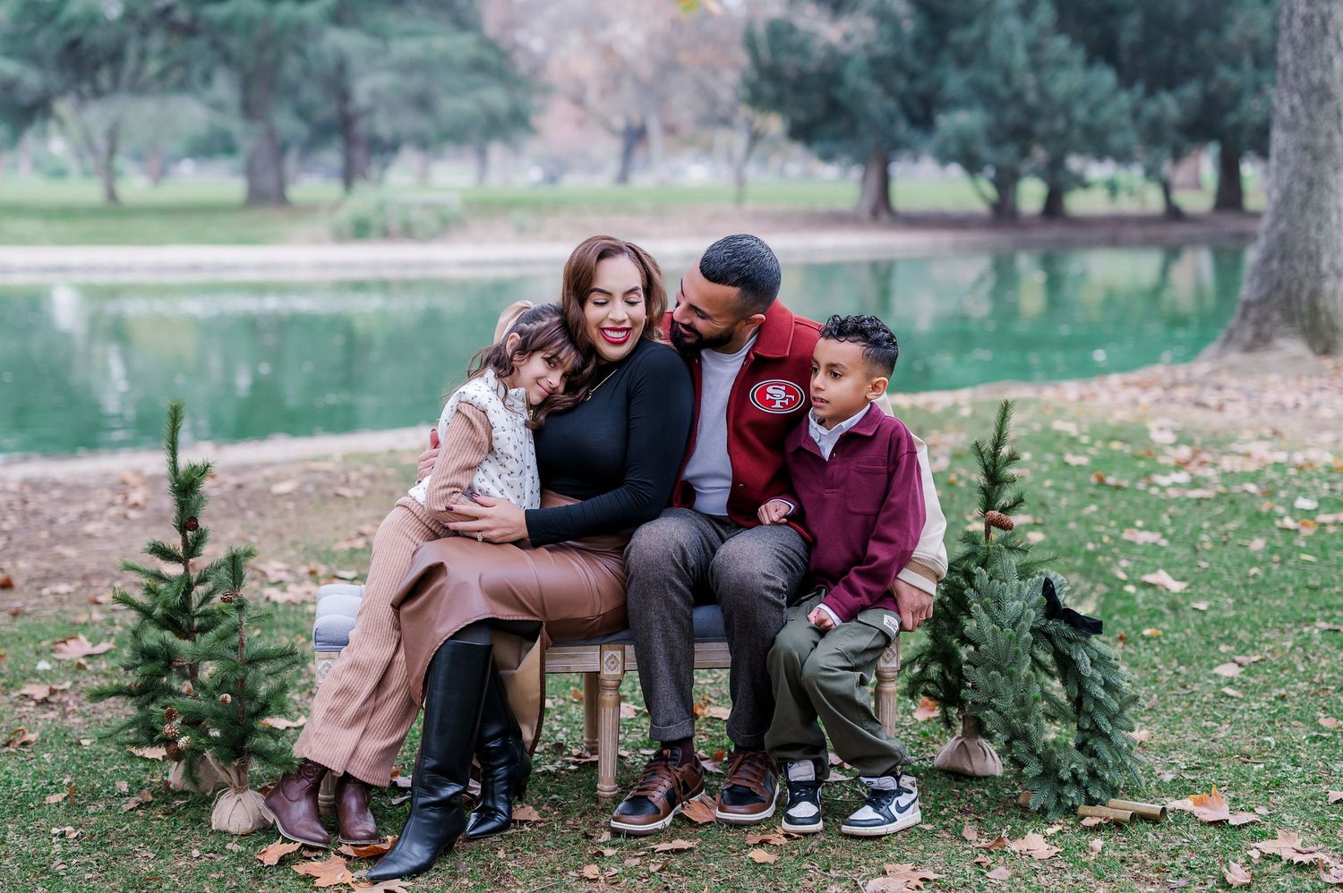 A couple sits together on a park bench surrounded by autumn leaves and evergreen trees in a romantic outdoor setting.