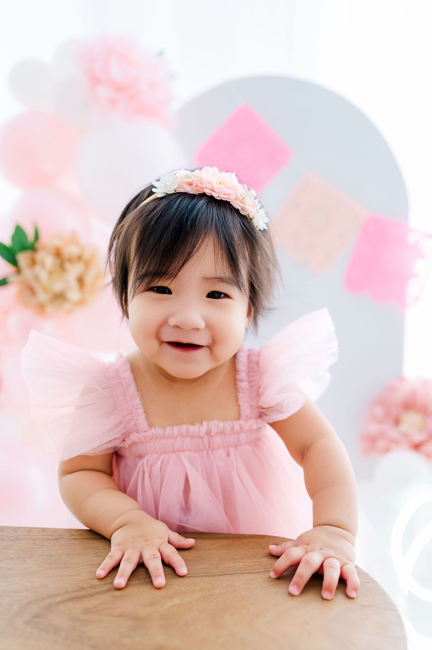 Smiling baby girl in pink tutu dress and floral headband at first birthday party with pink balloon decorations.