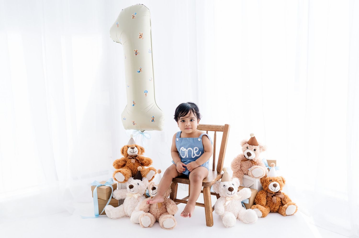A toddler sits on a chair surrounded by teddy bears next to a large number one balloon for a first birthday photo.