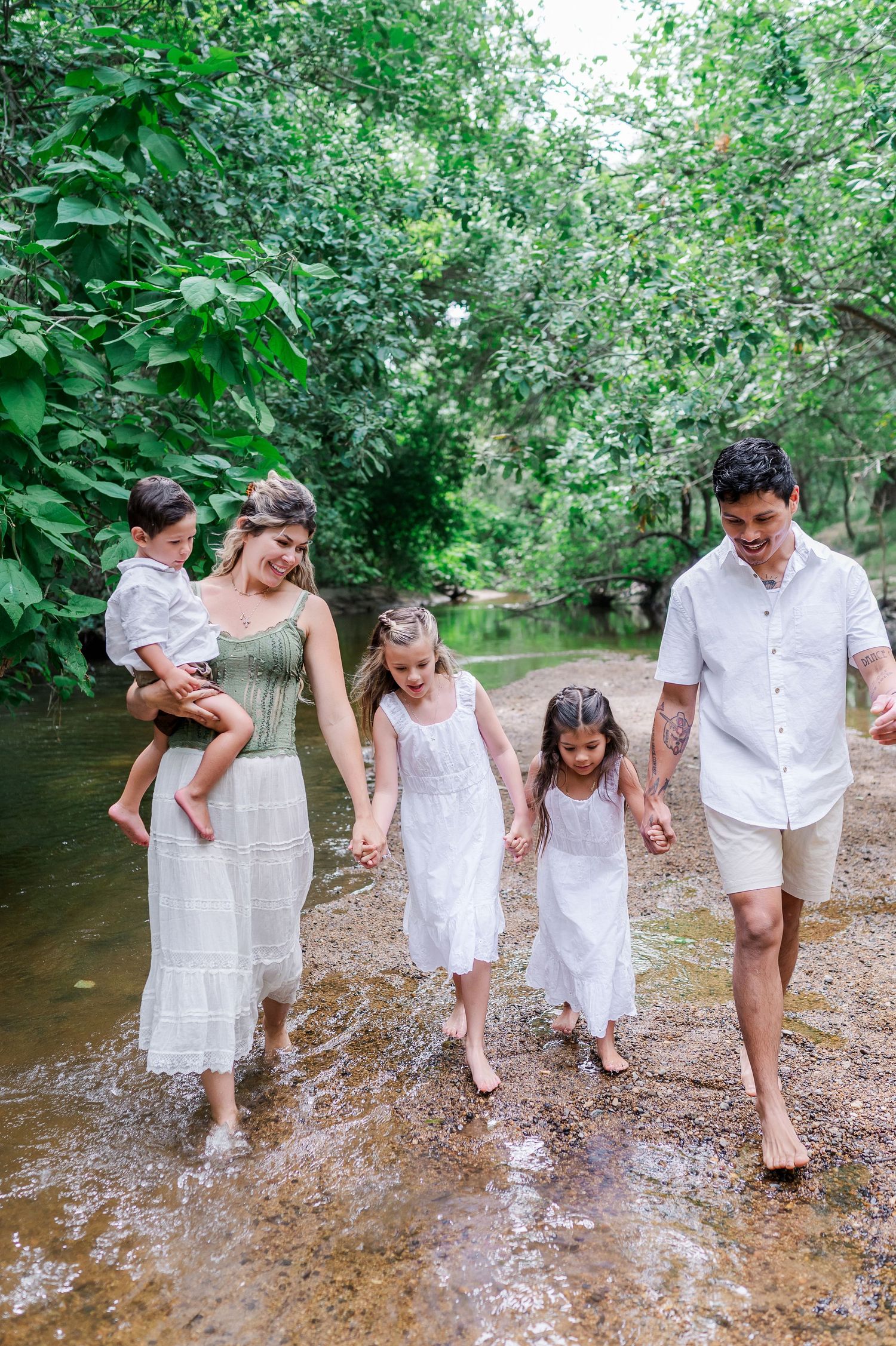 Family of five walking barefoot through a shallow creek surrounded by lush green trees, wearing white and green outfits.