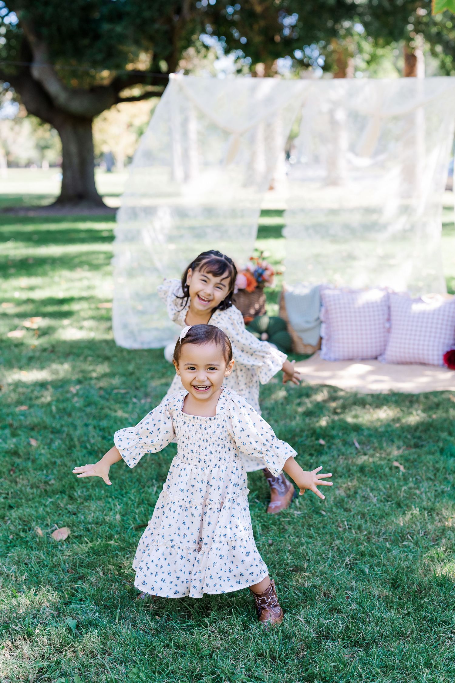 A small child wearing a white polka dot dress plays and twirls joyfully on green grass in a sunny outdoor setting.