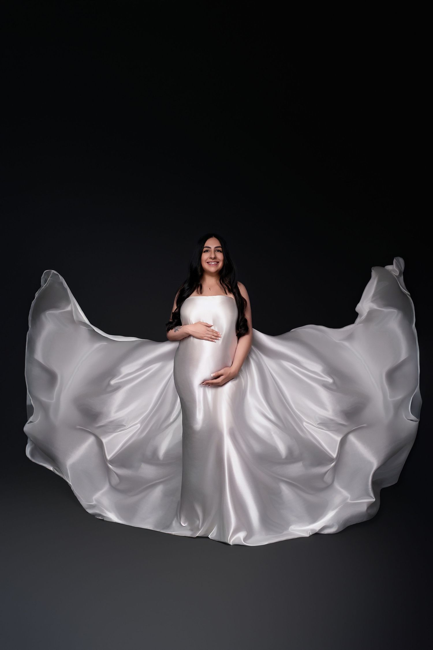 Dramatic maternity portrait featuring flowing white dress against dark backdrop.