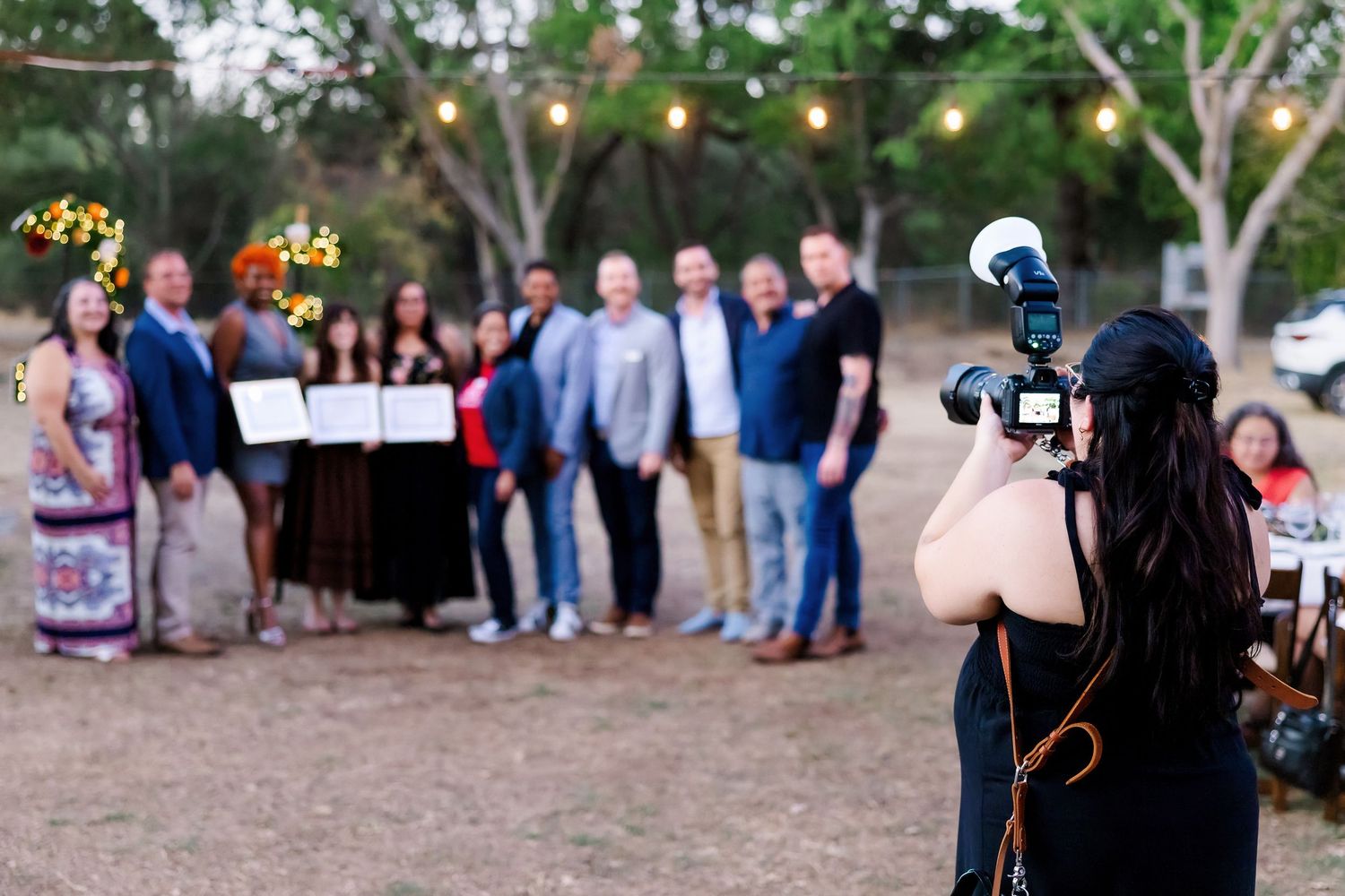 A photographer capturing a group photo of wedding guests at an outdoor venue with string lights.
