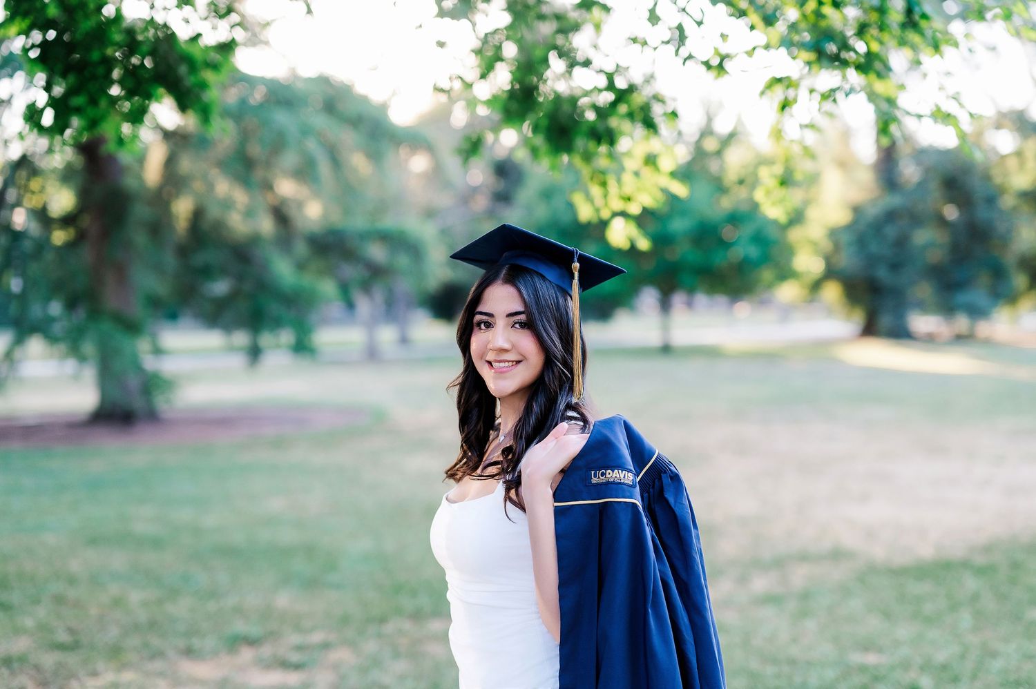 Graduate in academic regalia walking through a tree-lined path with string lights overhead.