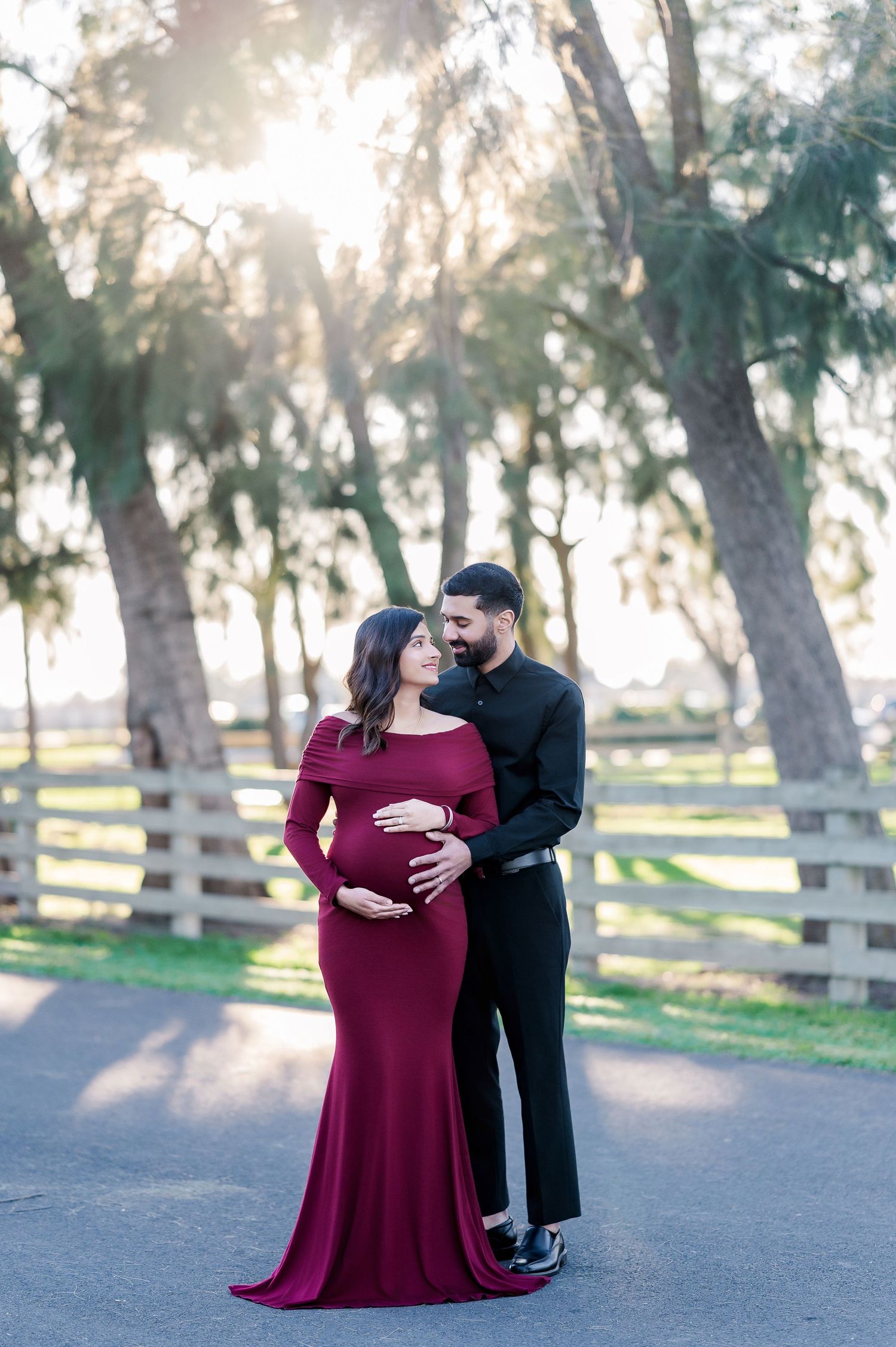 Couple's maternity session in burgundy dress and black outfit against rustic park setting at sunset.