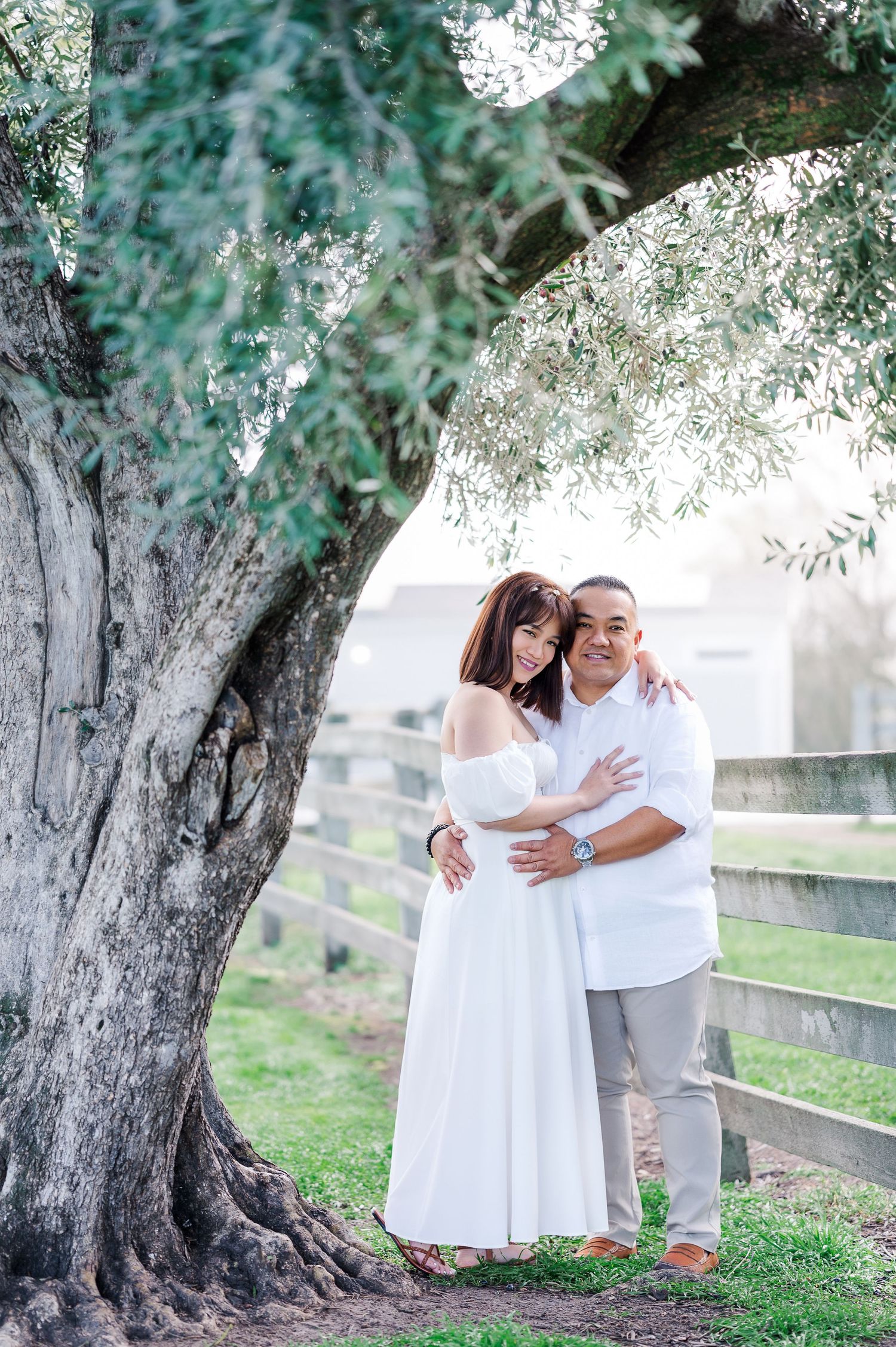 A couple embraces under an olive tree in white outfits during an outdoor photoshoot.