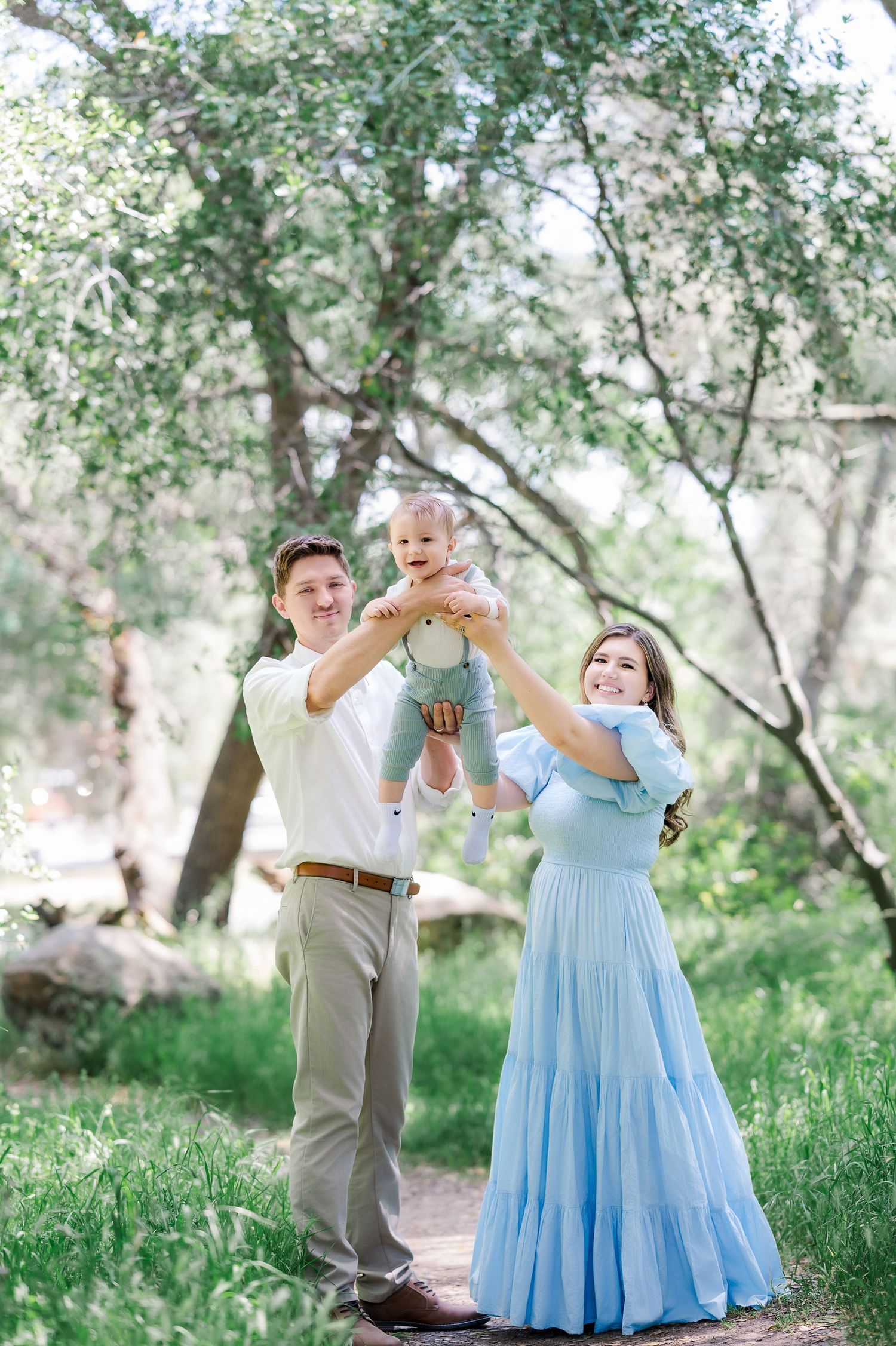 A couple in formal attire dances together in a sunlit garden setting with trees and greenery in the background.