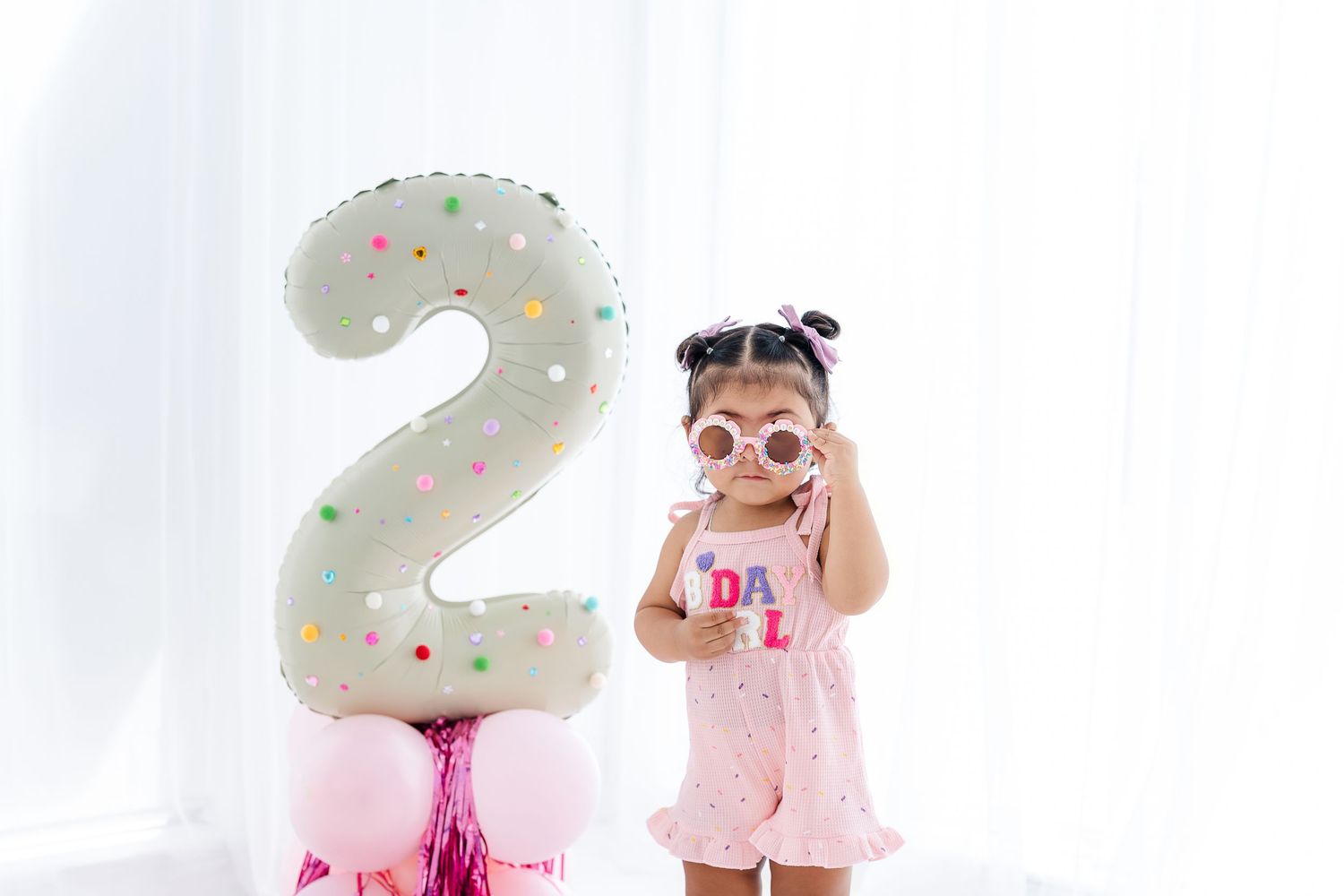 Toddler girl in pink outfit holds sunglasses beside a large number 2 balloon for her 2nd birthday celebration.