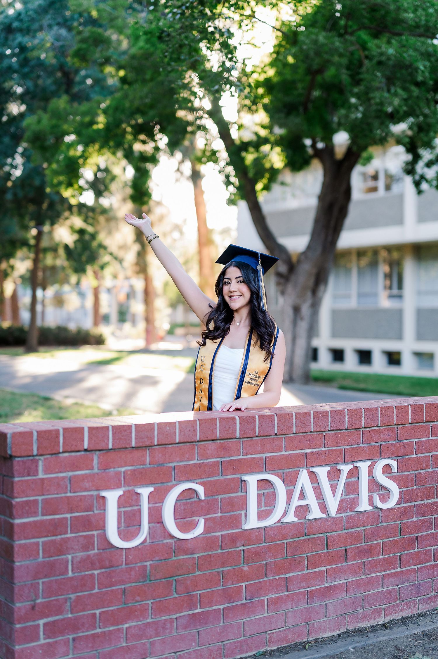 Graduate in academic regalia posing by the UC Davis brick sign wall on campus.