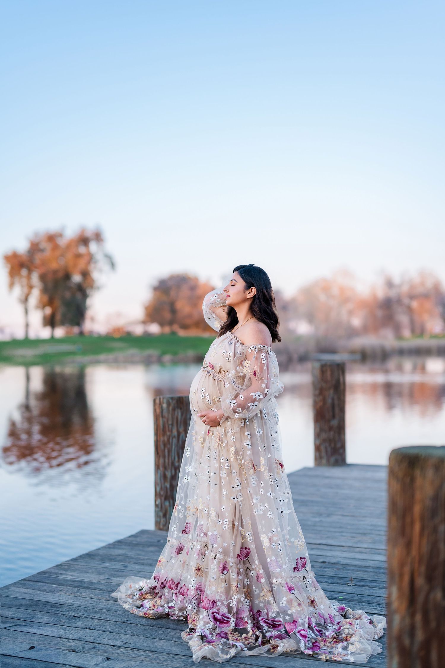 A flowing floral dress maternity session on a wooden dock overlooking a serene lake at sunset.