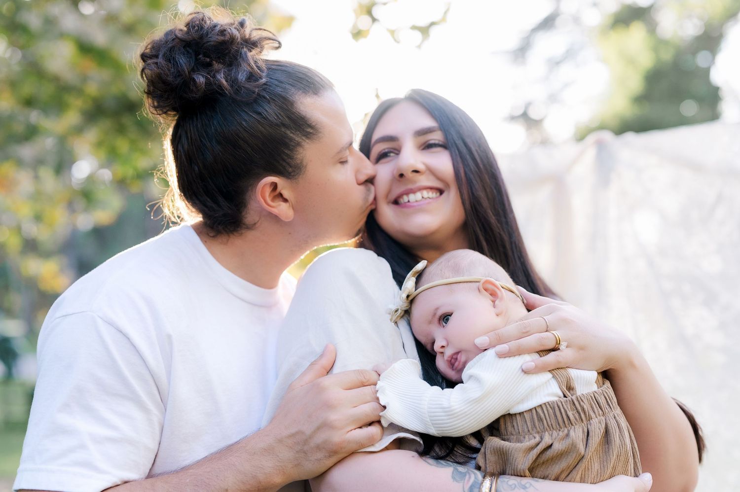 A happy couple kisses and cuddles while holding their newborn baby outdoors in a sunlit park setting.