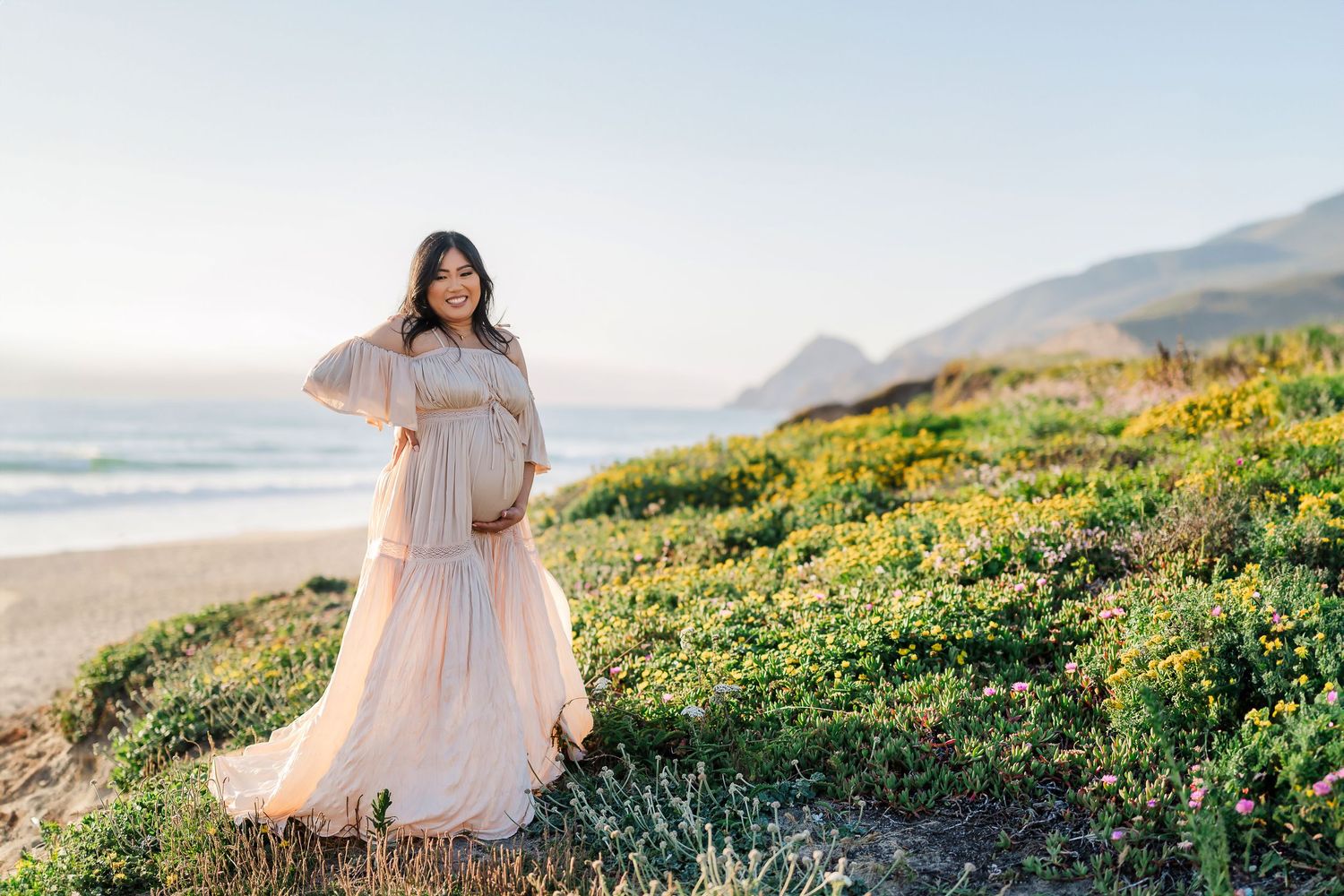Pregnant woman in flowing blush gown poses on coastal bluff with ocean waves and golden hills behind her.
