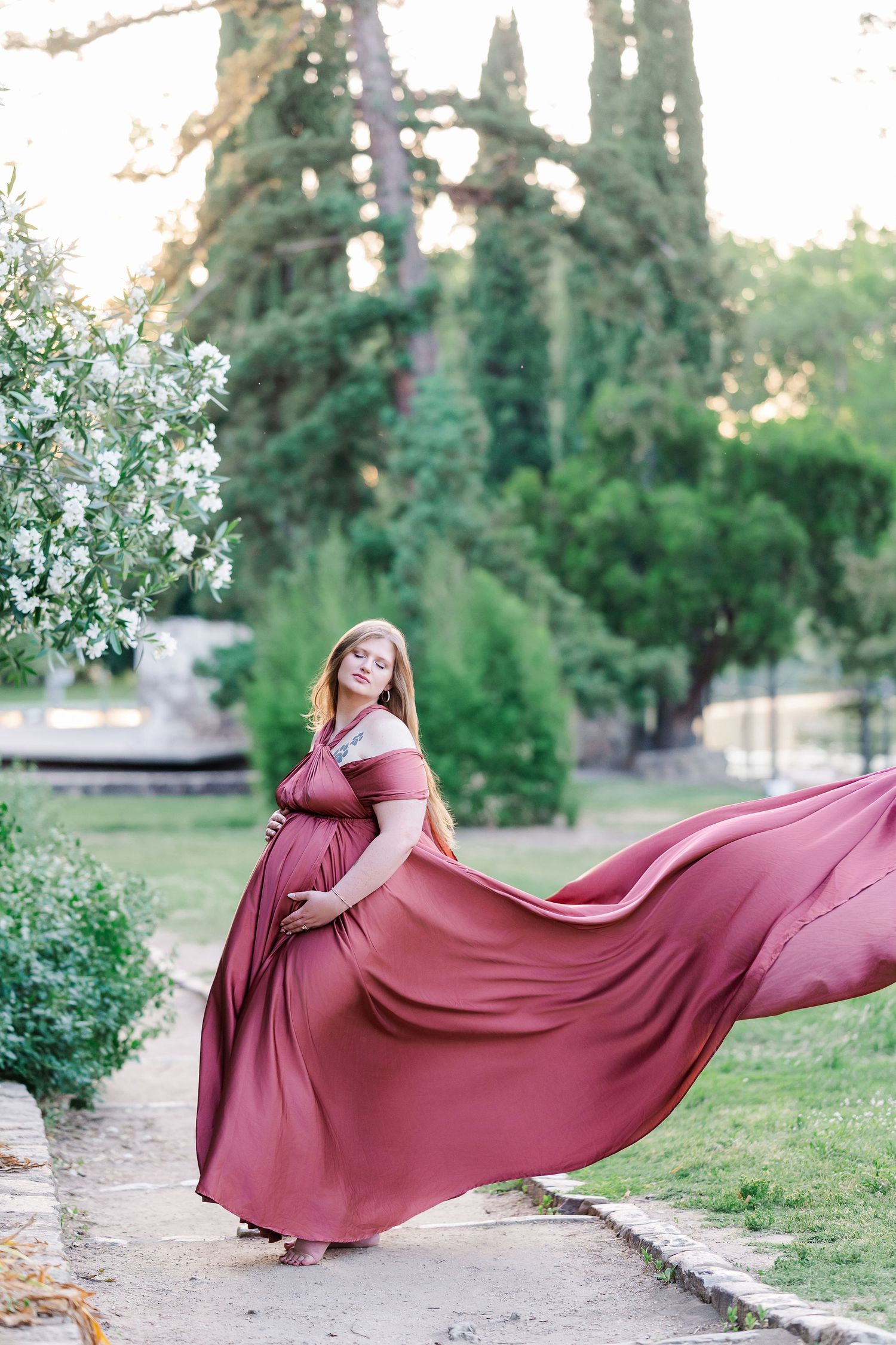 Pregnant woman in flowing dusty rose gown poses gracefully in lush garden, fabric billowing in the breeze. Sacramento Maternity Photographer