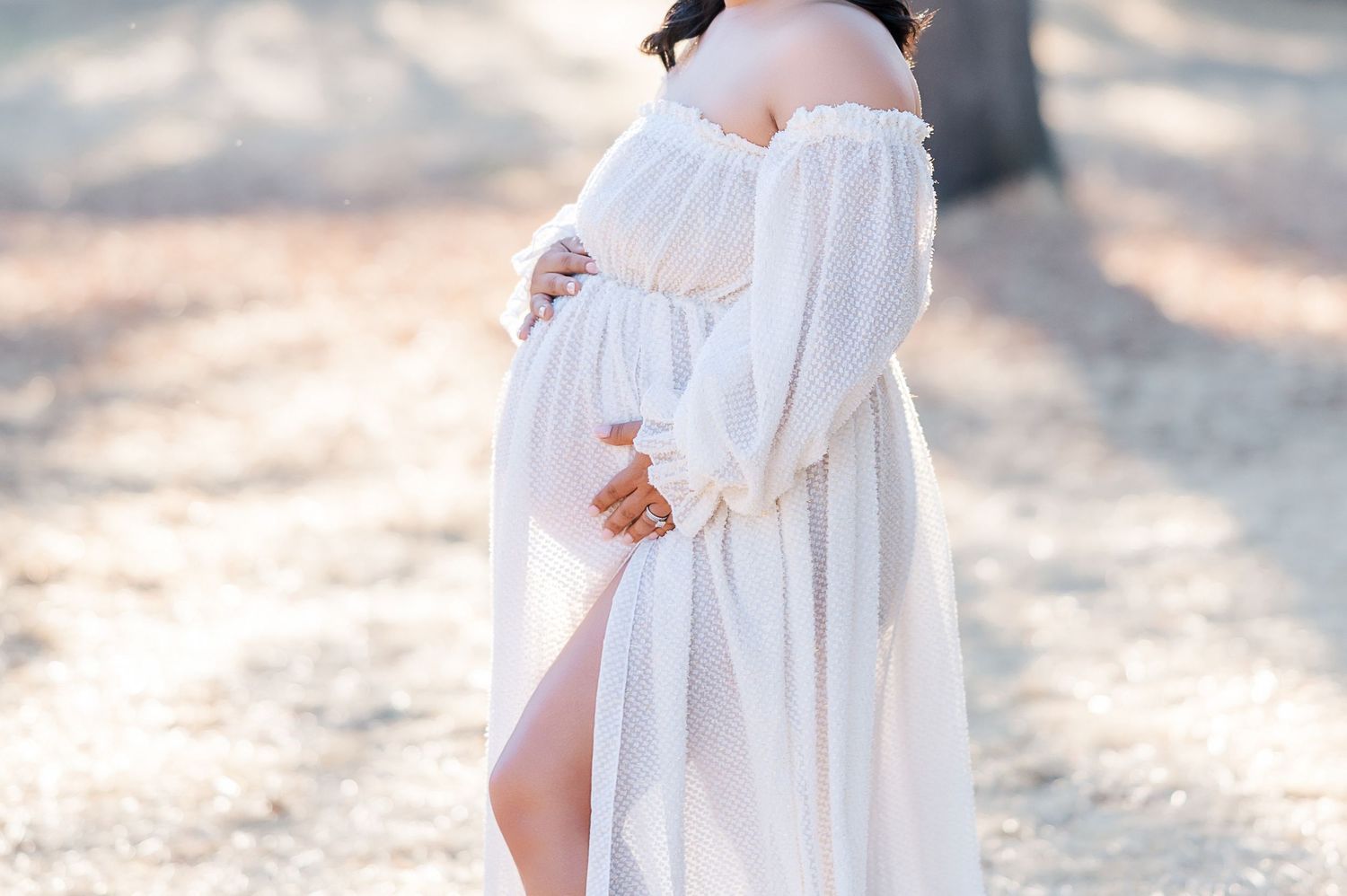 Pregnant woman in white off-shoulder flowing gown cradling baby bump outdoors in soft natural light.