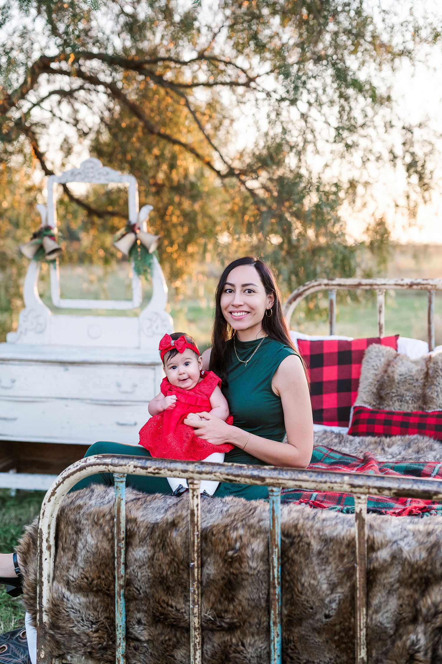 A woman in a green dress holds a baby in a red outfit on a vintage iron bed with holiday decor outdoors.