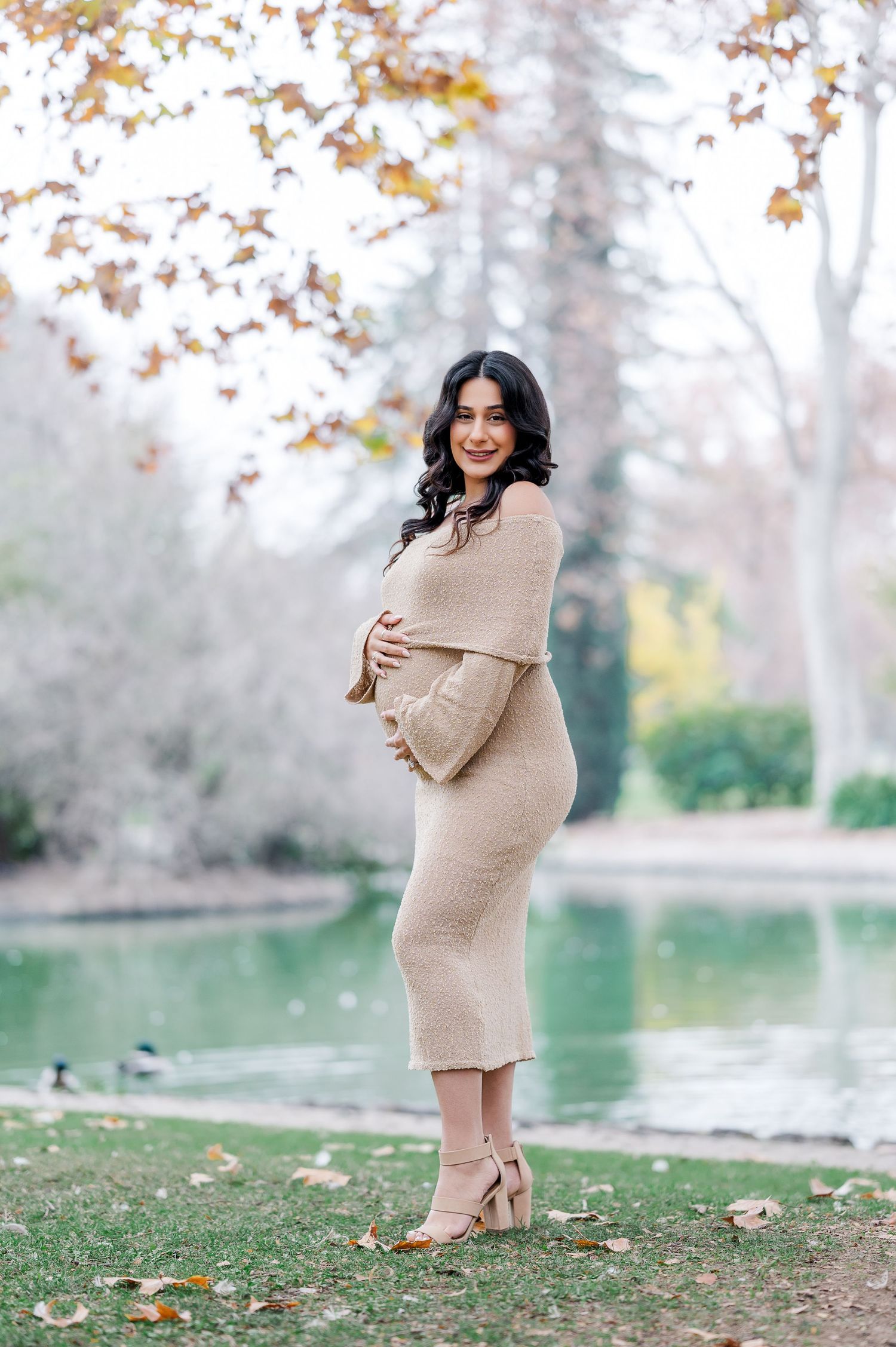 A maternity photo session in a park during autumn shows a woman in a beige sweater dress by a lake.