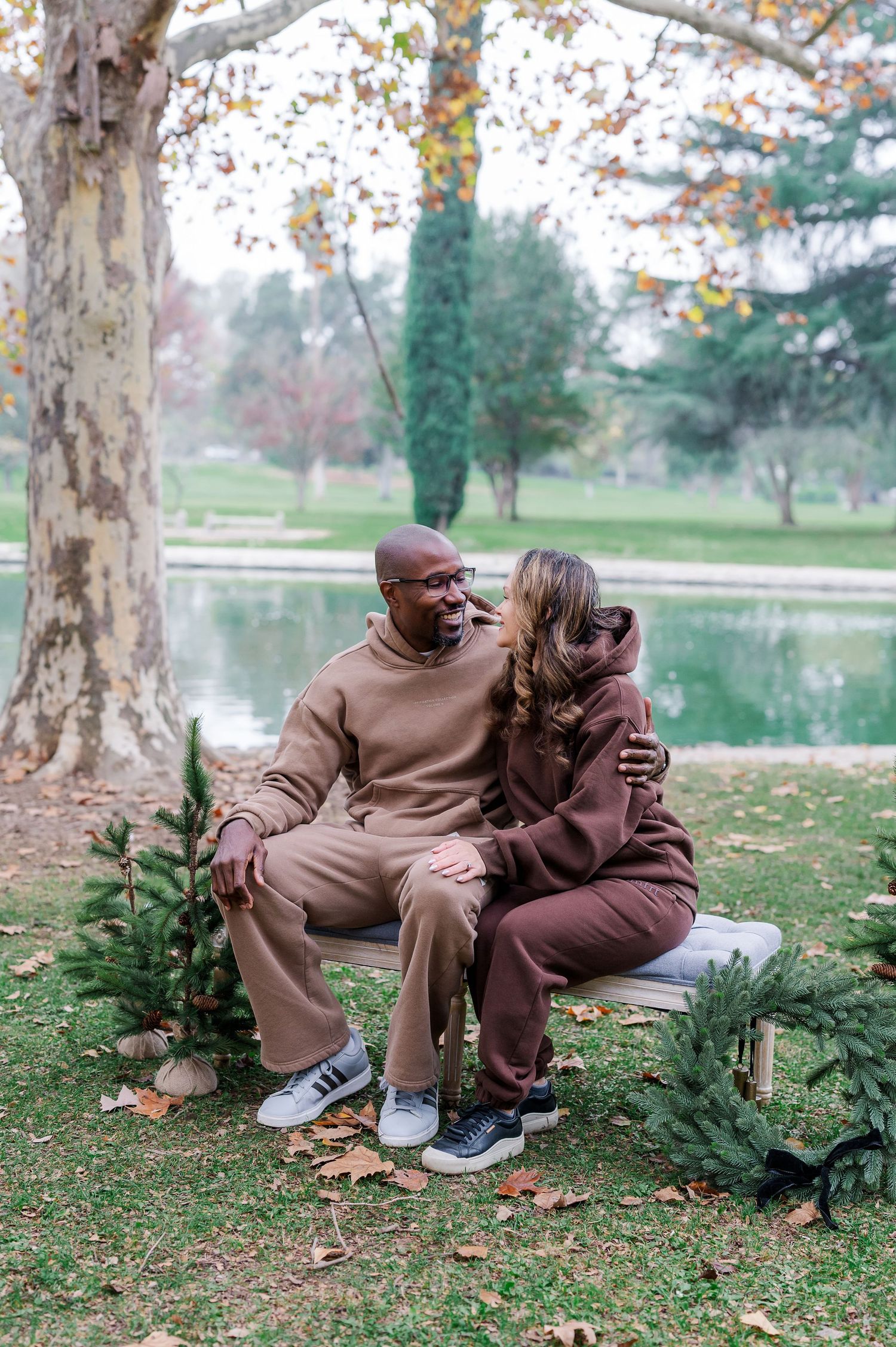 A couple sits together on grass near evergreen bushes in a park on an autumn day wearing matching brown outfits.