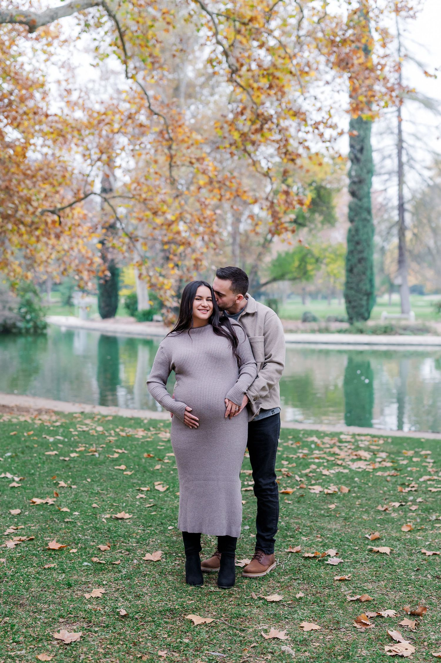 A couple embraces near a tranquil pond surrounded by autumn trees with golden leaves scattered on the grass.