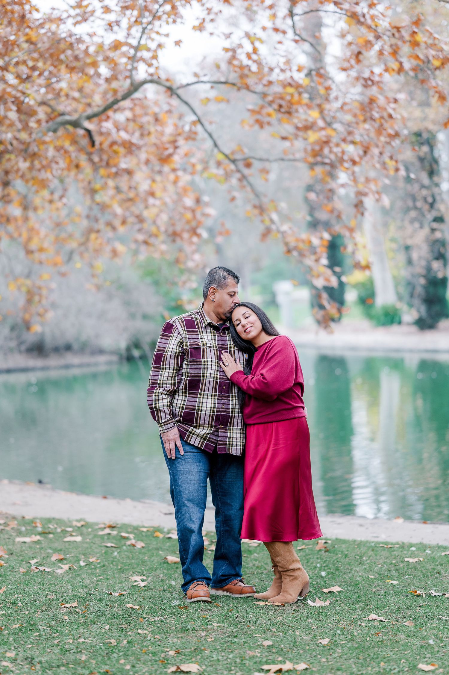 A couple embraces by a lake in autumn, with orange leaves overhead and water reflecting the fall scenery.