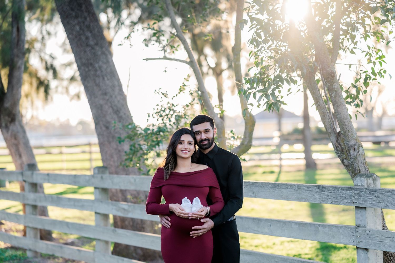Maternity photography session in a sun-dappled park with wooden fences and tall trees.