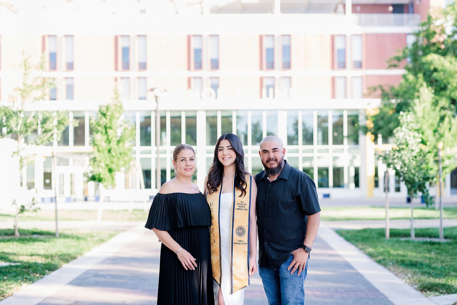 Three people stand together in graduation attire on a university campus walkway with brick buildings in background.