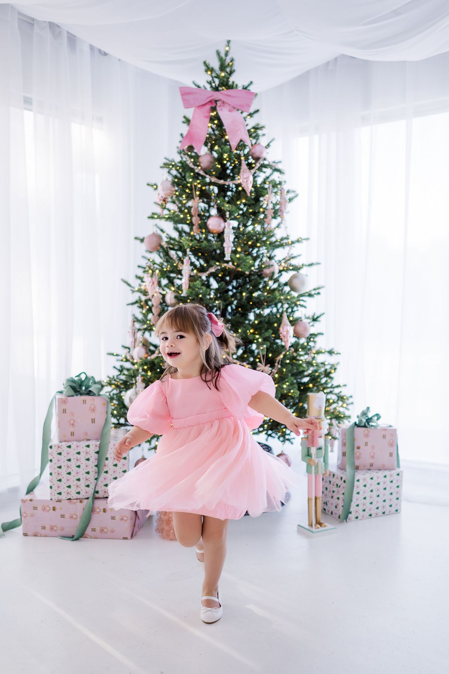 A child in a pink tulle dress twirls joyfully in front of a Christmas tree with silver and pink wrapped presents.