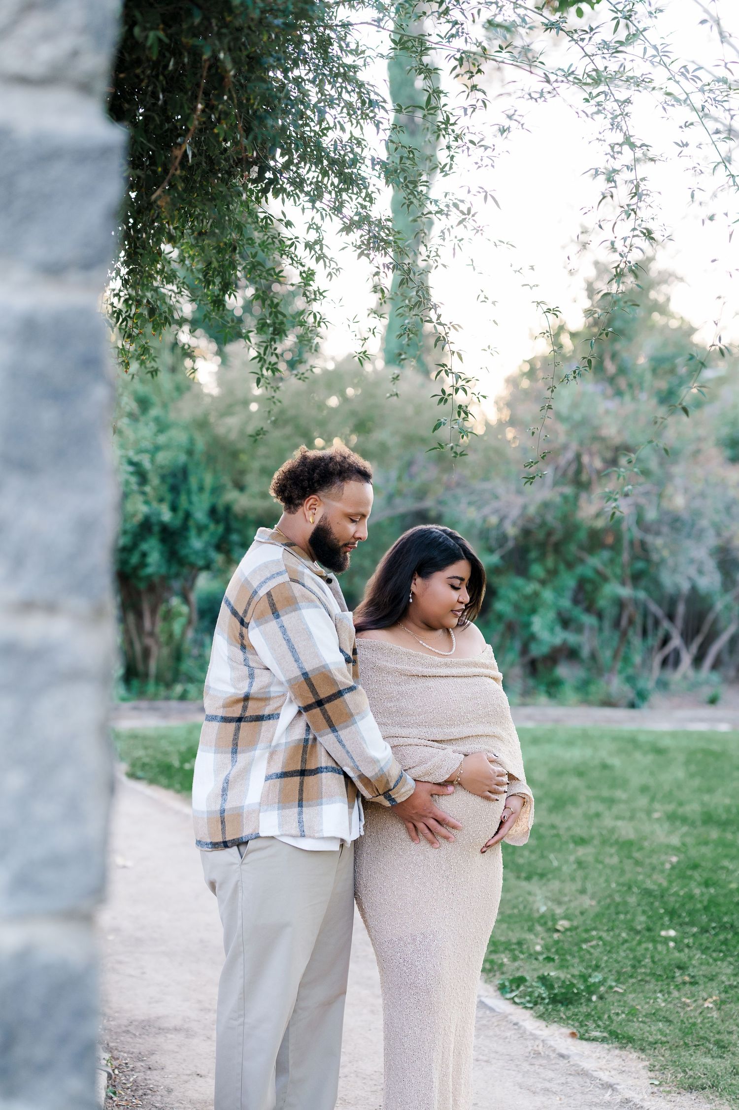 A couple shares an intimate moment on a garden path, with the person in beige embracing their pregnant partner.