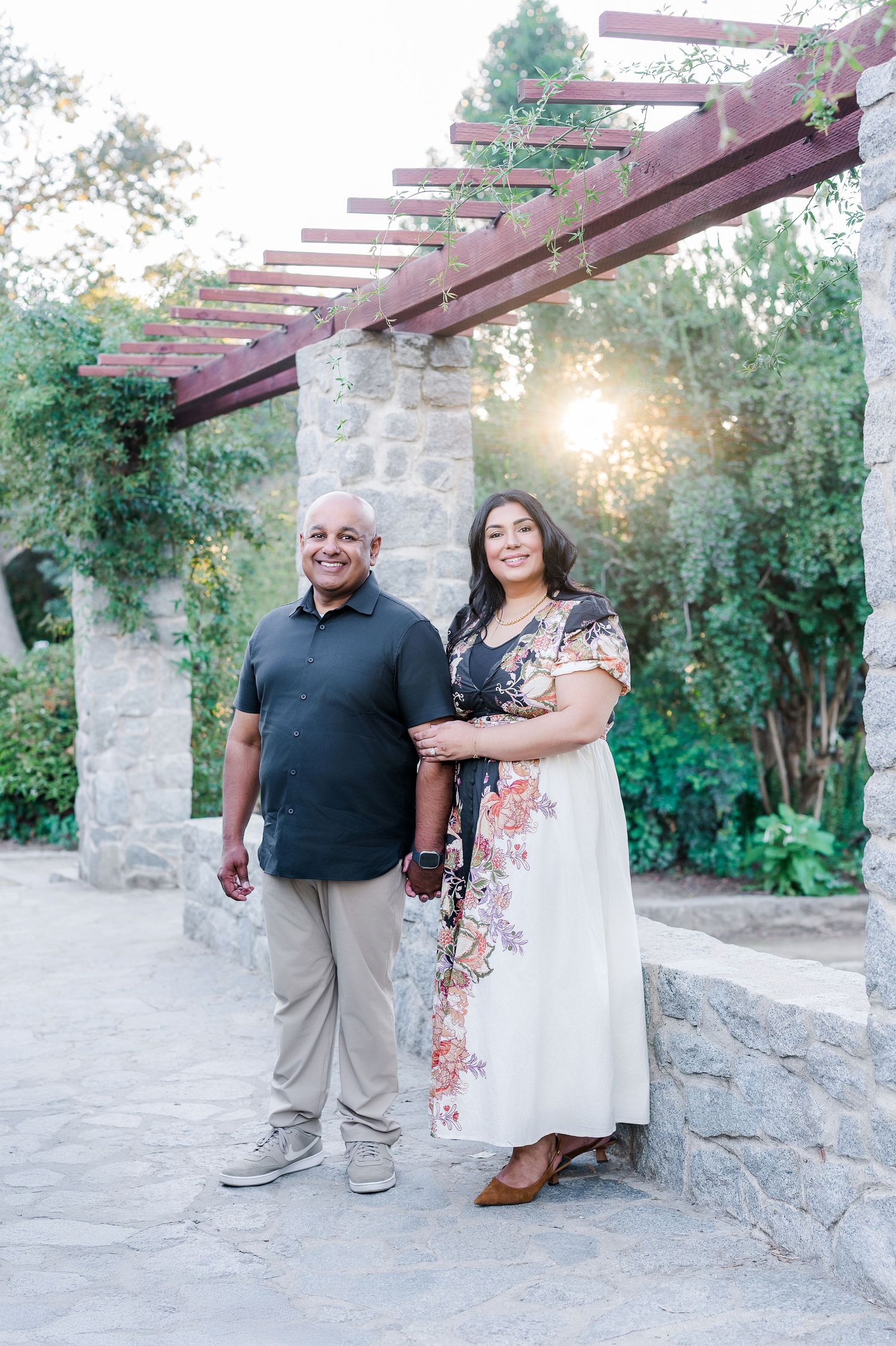 A couple stands together under a wooden pergola in a garden setting during golden hour.
