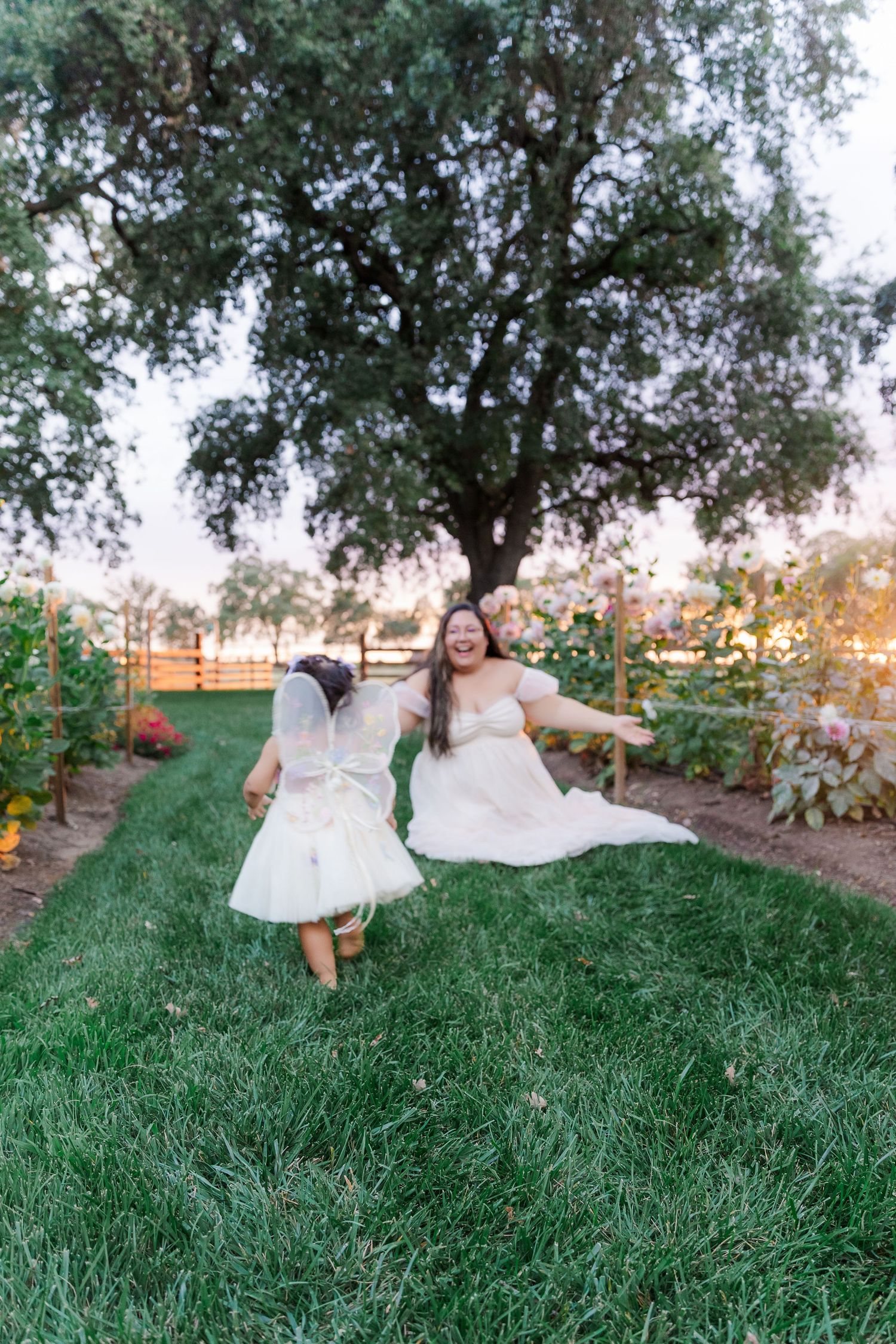 Bride in white gown laughing and playing with flower girl in garden at golden hour sunset.