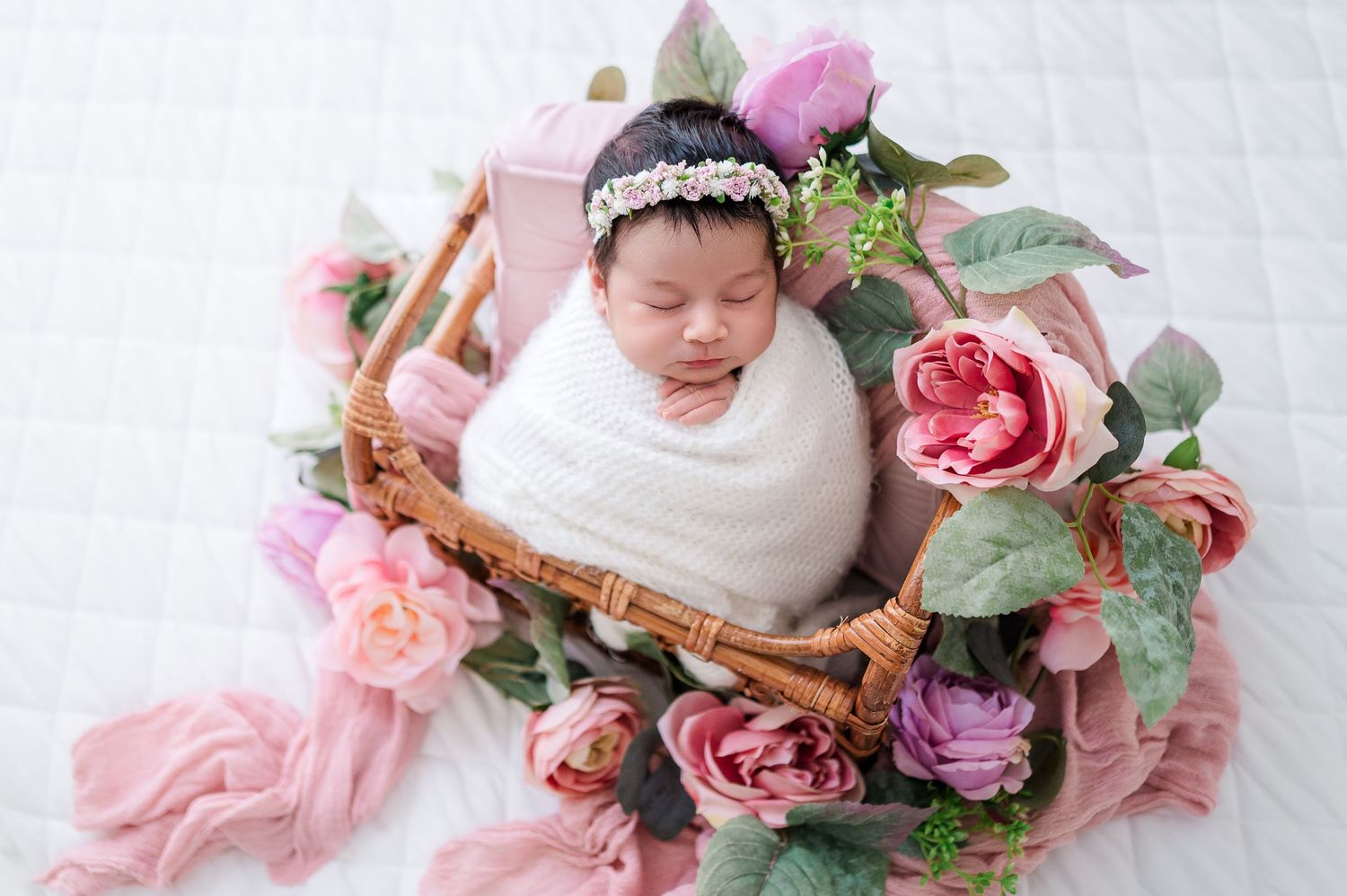 Newborn baby swaddled in white wrap sleeping peacefully in a wicker basket surrounded by pink and purple flowers. Sacramento Newborn Photo