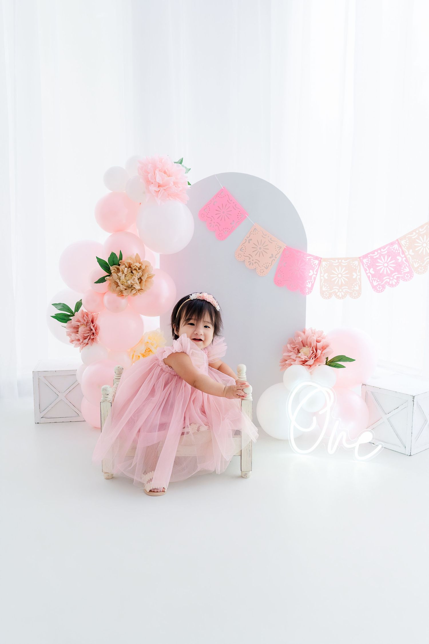A toddler girl in a pink tulle dress celebrates her first birthday with balloon and floral decorations.