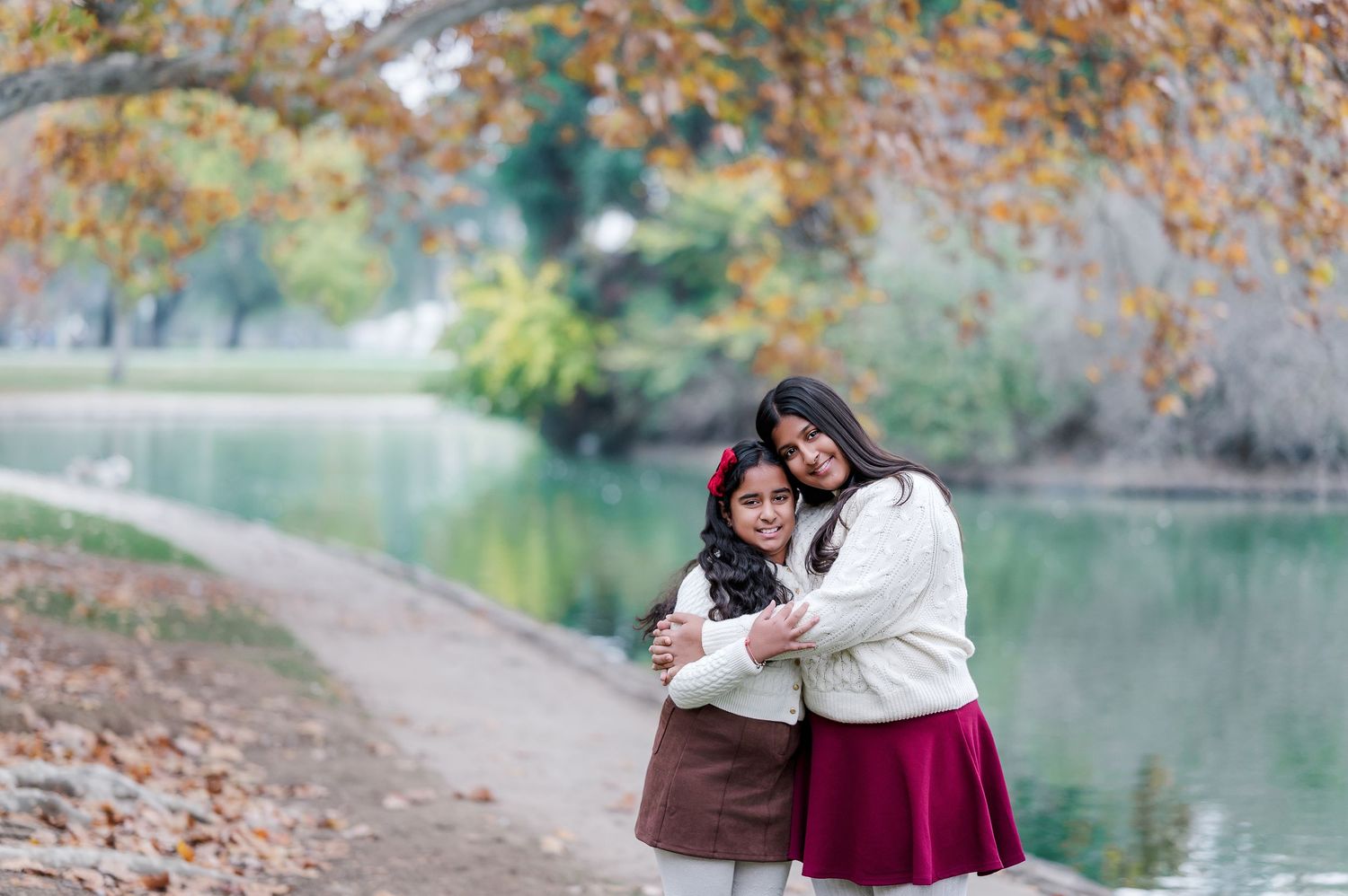 Two women hugging warmly by a scenic autumn park lake, surrounded by colorful fall foliage.