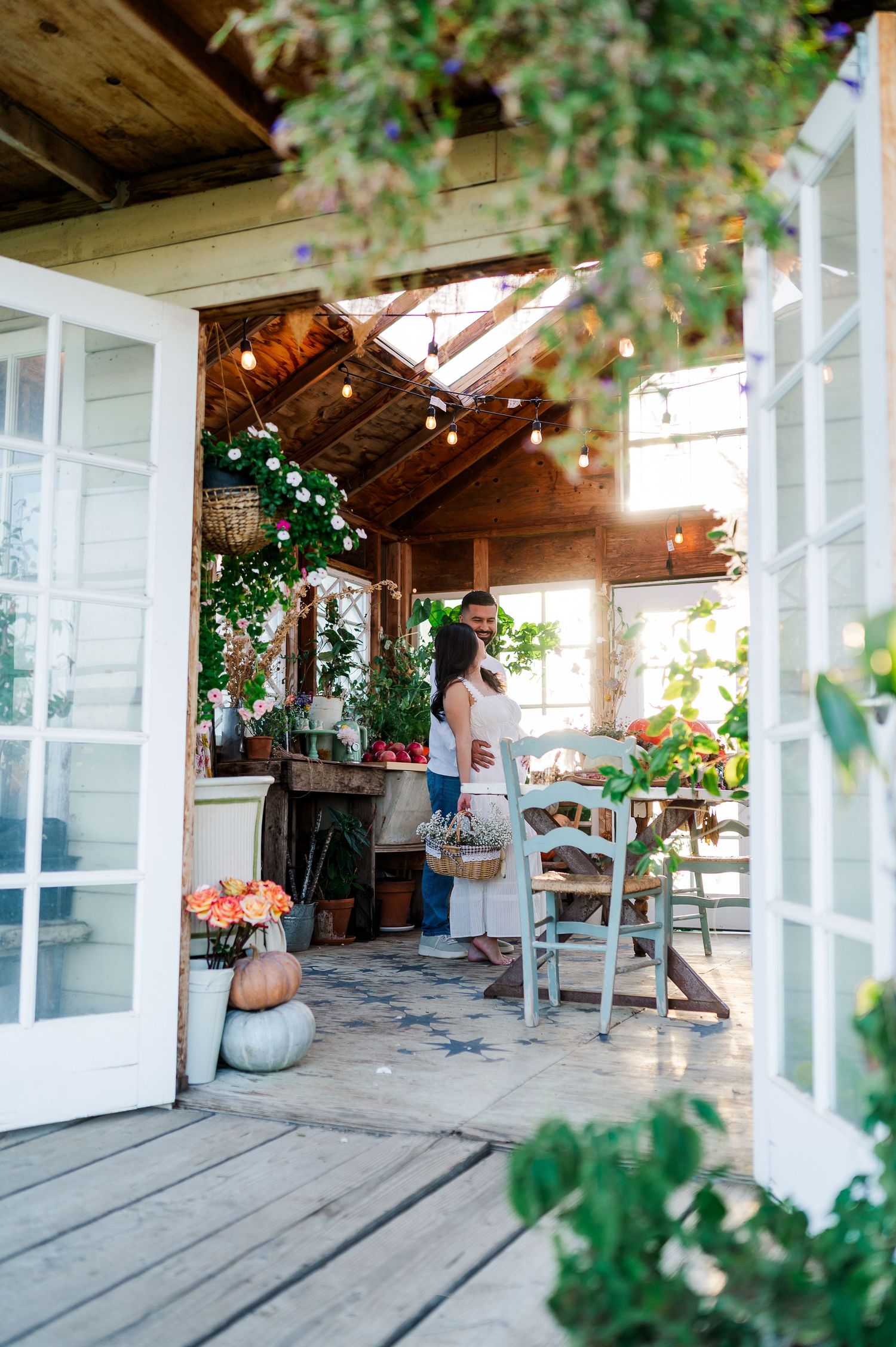 Couple embracing in a charming rustic greenhouse with string lights, flowers, and pumpkins on a wooden porch.