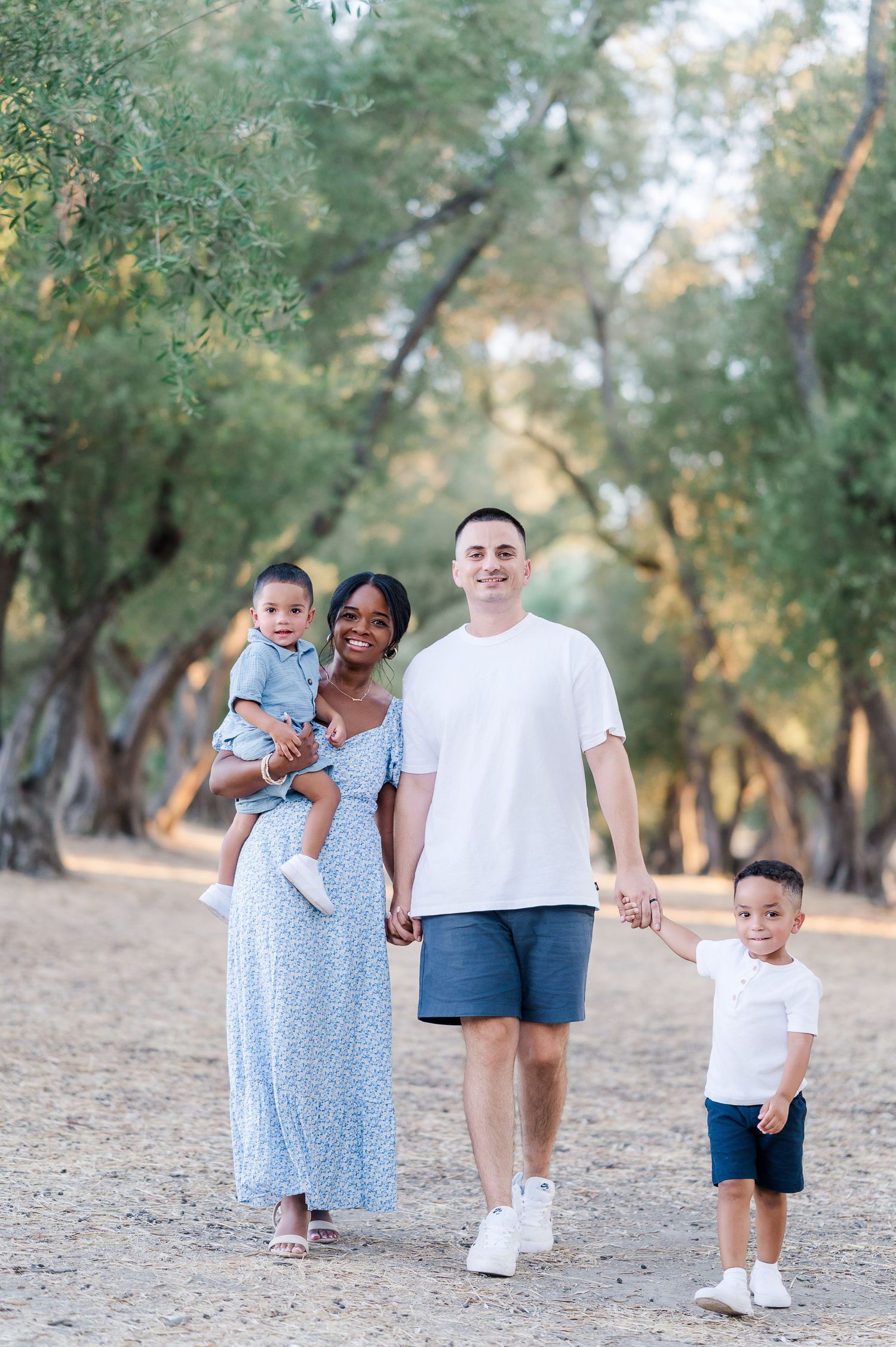 Happy interracial family of four walking outdoors among green trees, parents holding children hands on a sunny day.
