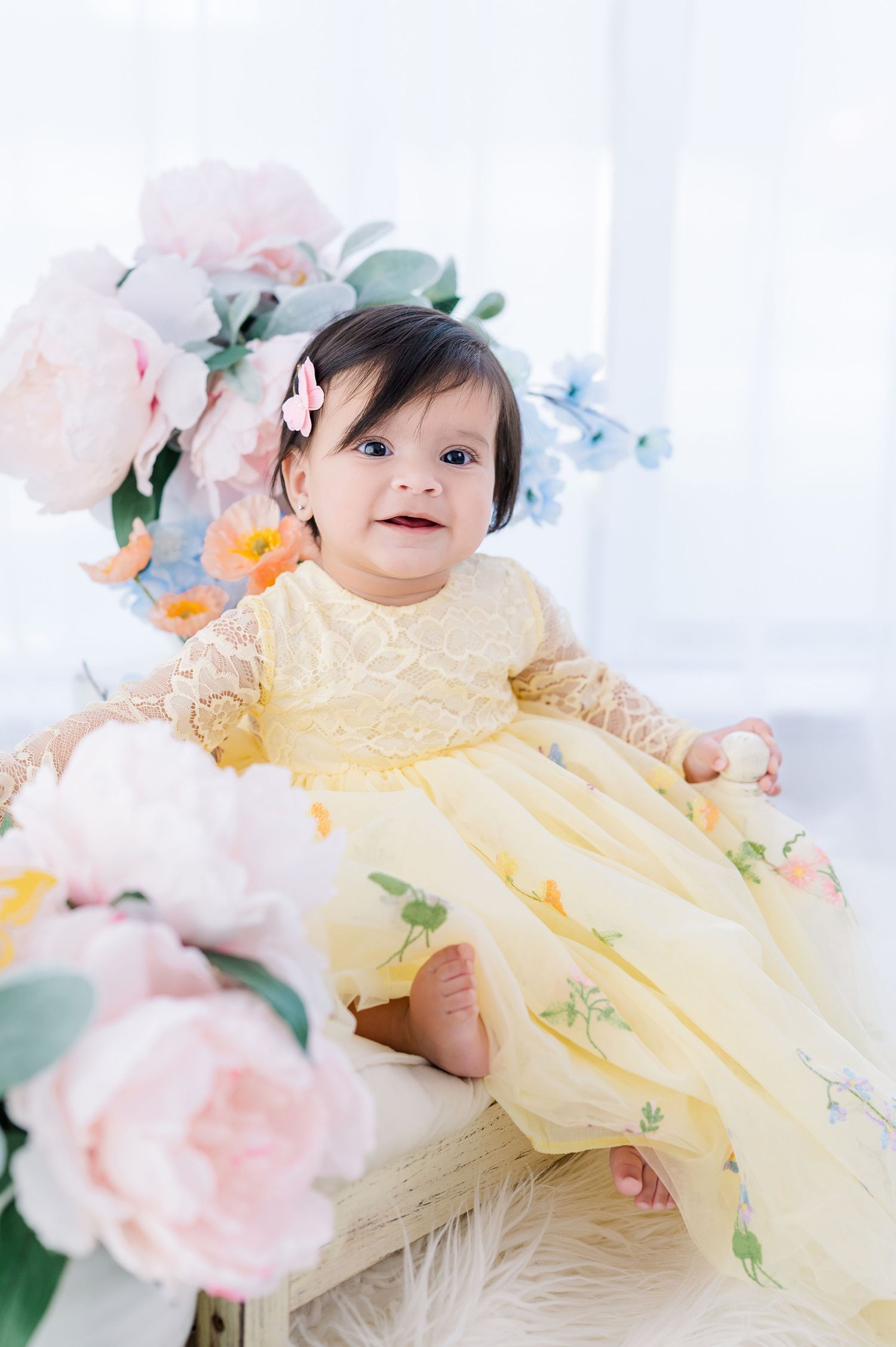 A smiling baby girl in a yellow lace dress sits among pink and blue flowers in a bright studio setting.