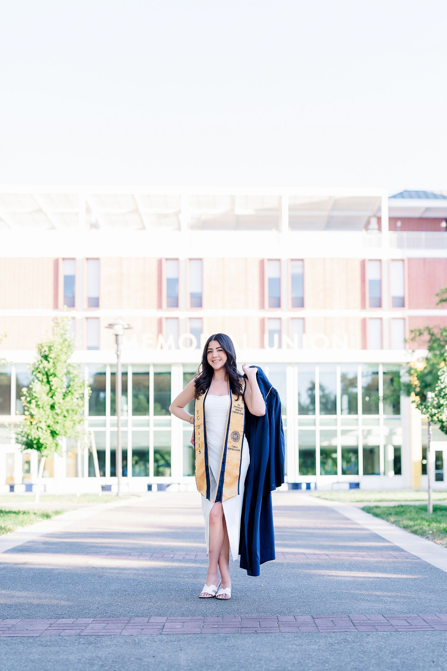 Graduate in cap and gown walking across an open plaza on the UC Davis campus.