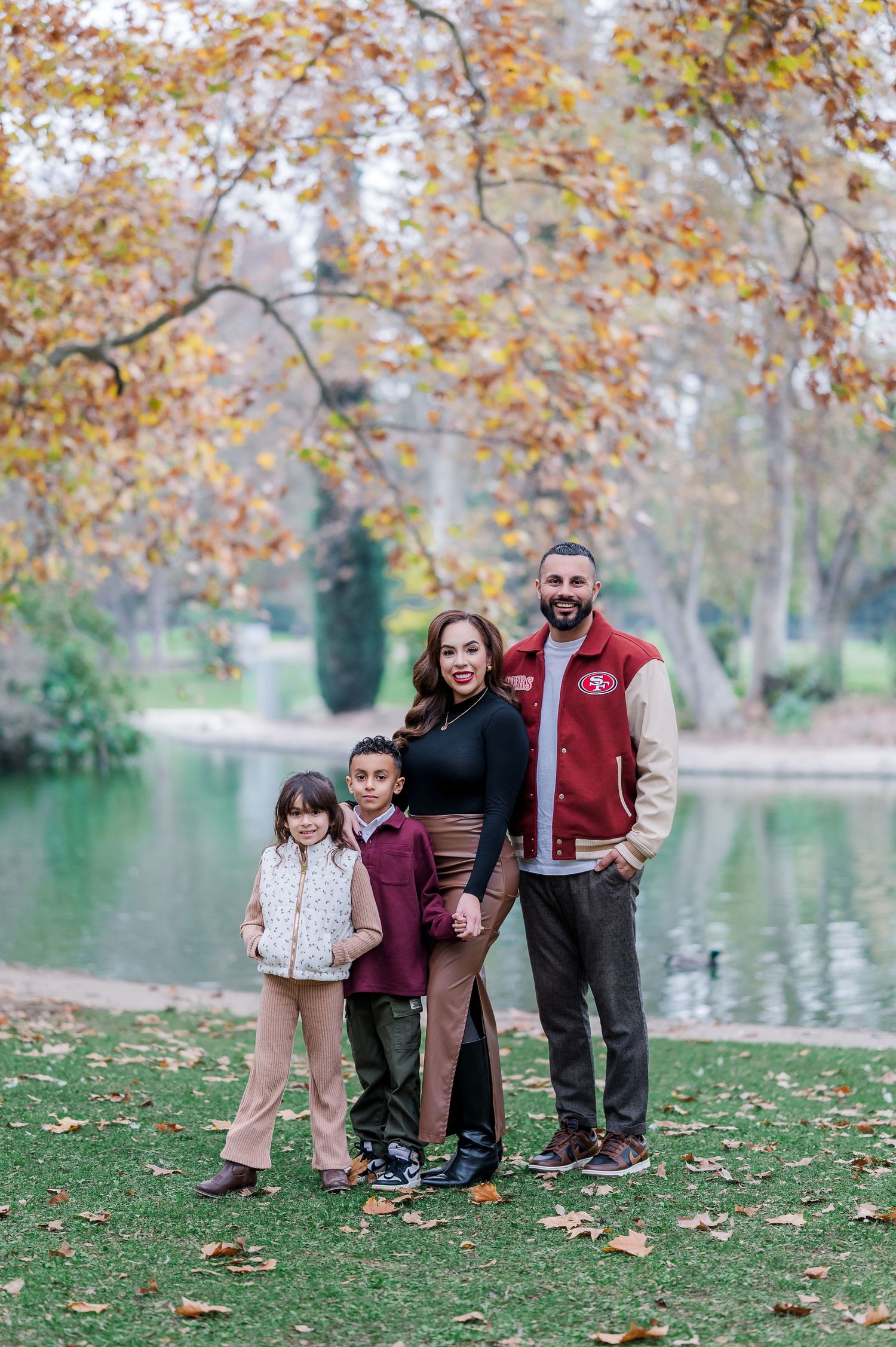 A family stands together on grass near a tranquil pond surrounded by autumn trees with golden leaves.