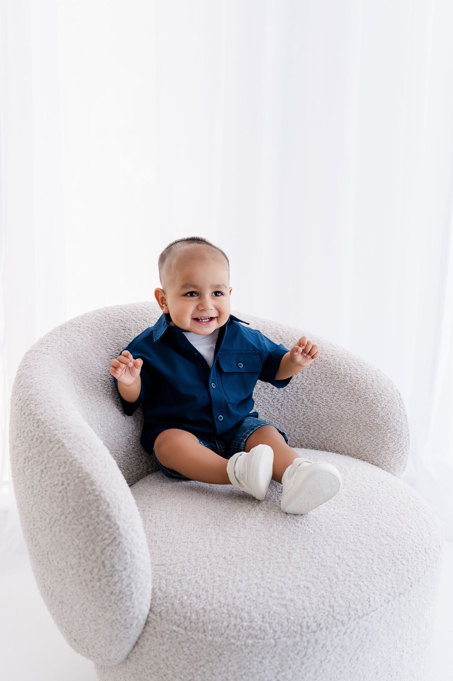 Smiling baby boy in navy shirt sits in a white boucle chair, raising hands joyfully in a bright white room.