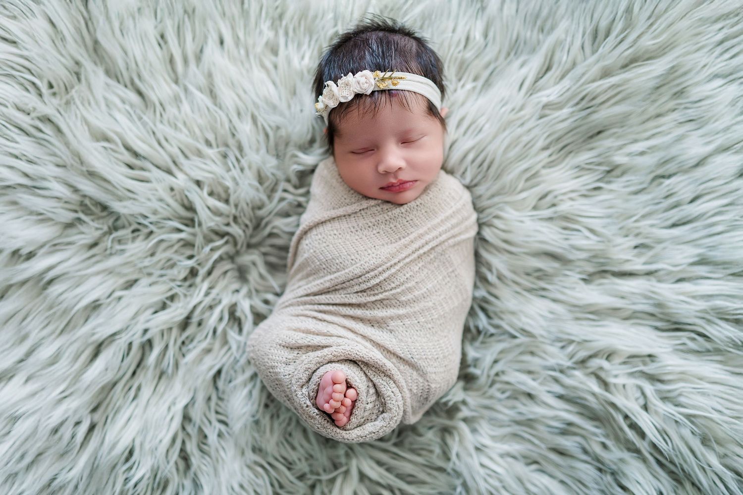 Sleeping newborn wrapped in beige swaddle on soft gray fur blanket wearing delicate headband.